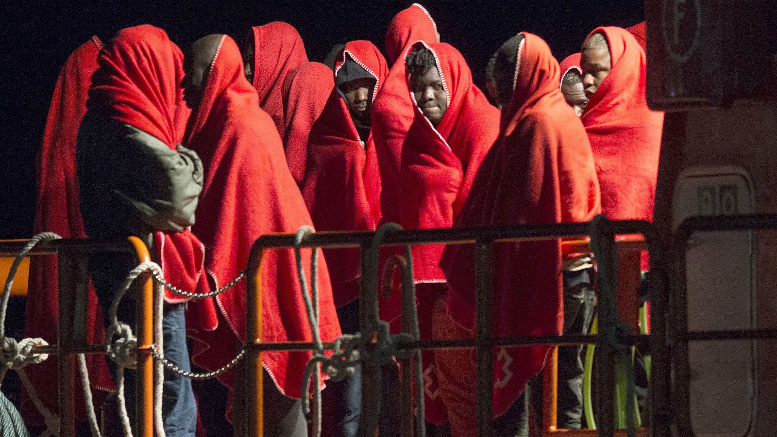 Inmigrantes abrigados con mantas rojas en el puerto de Almería tras ser rescatdos por Salvamento Marítimo de la patera en la que viajaban.