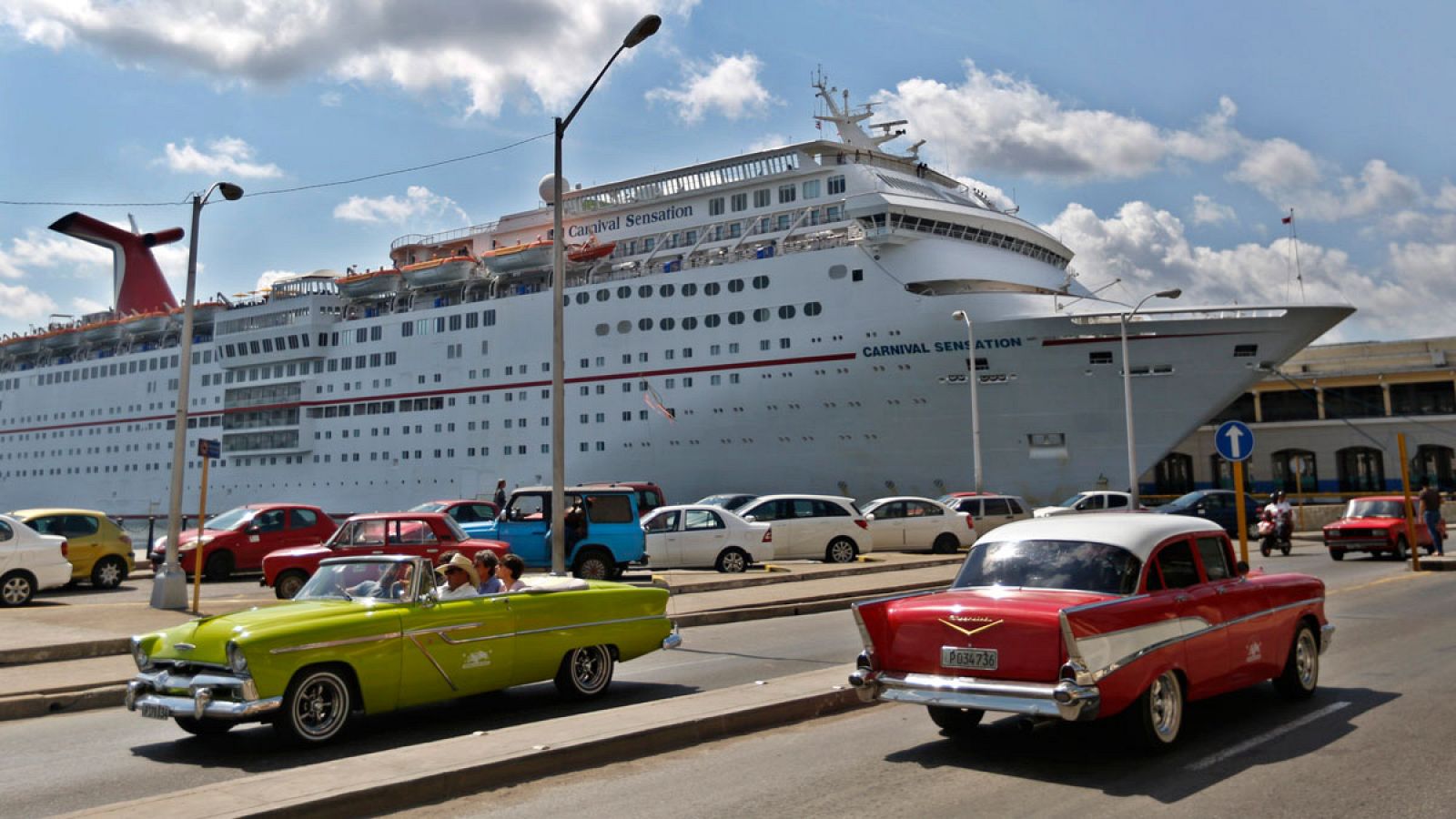 Dos coches estadounidenses clásicos pasan frente a un crucero anclado en el puerto de La Habana