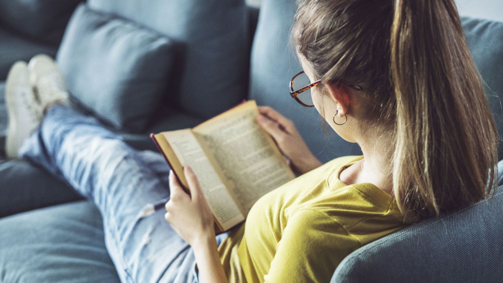 Una joven millennial lee una novela tumbada en el sofá.