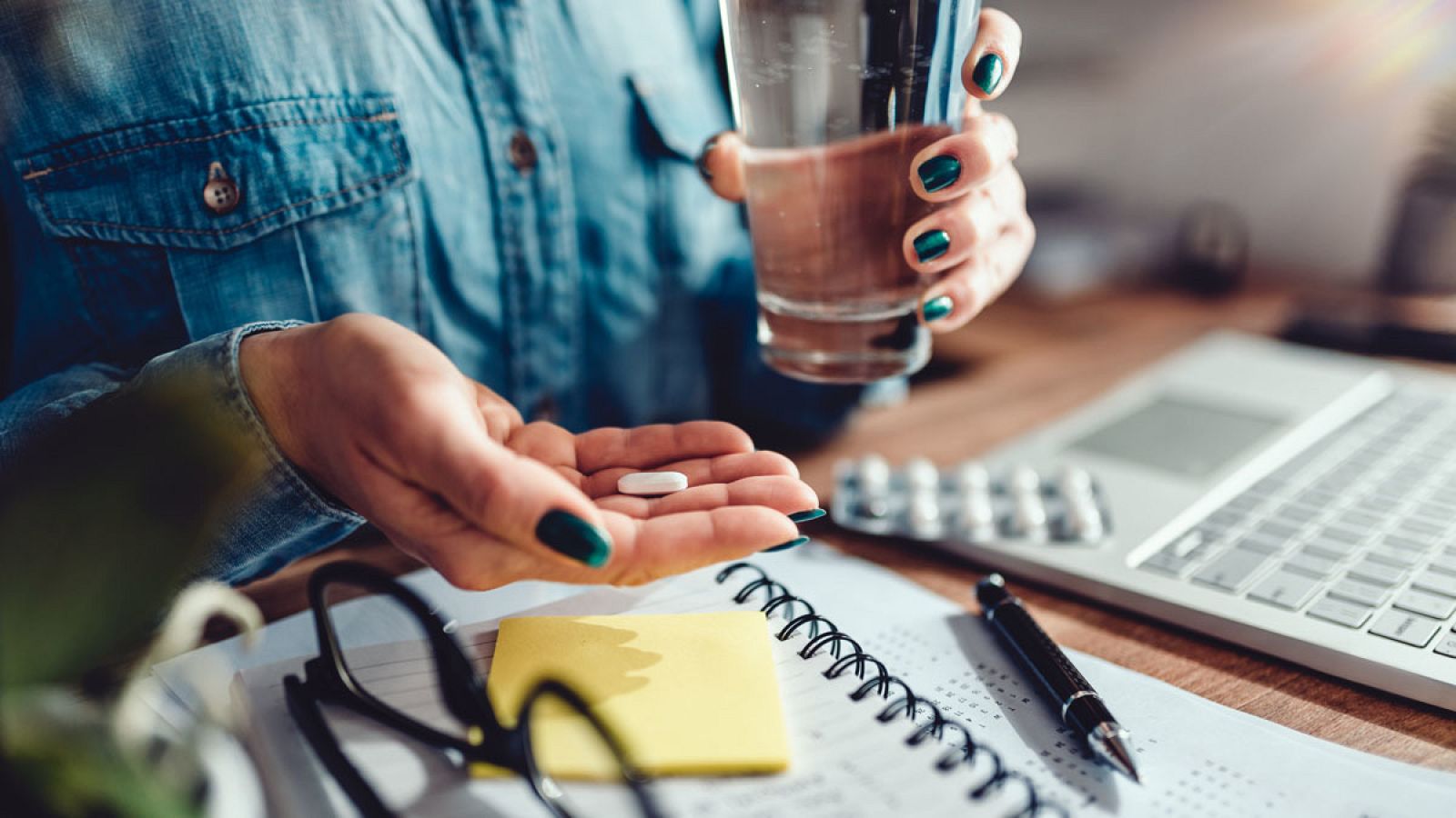 Una mujer se toma una pastilla, sentada en un escritorio de oficina, en una imagen de archivo