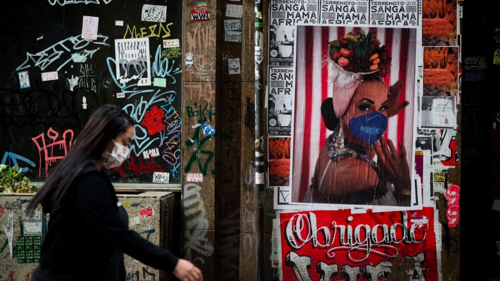 Una mujer con mascarilla caminando frente a un poster artístico en Sao Paulo.