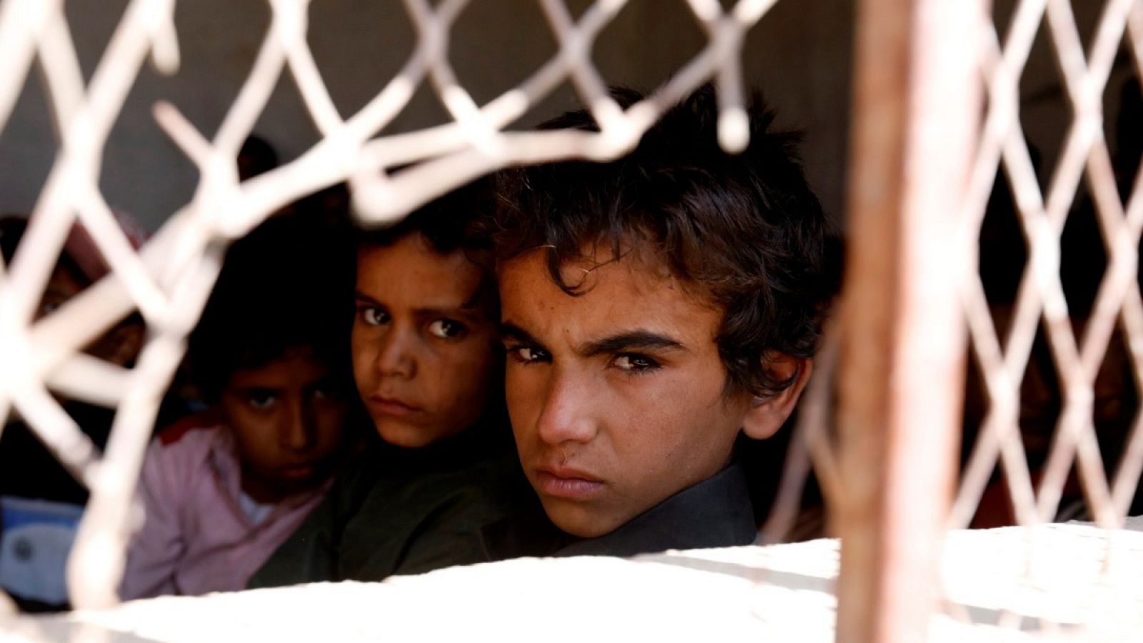 Tres niños yemeníes miran al cámara a través de la verja que cubre la ventana de su escuela, en Saná (Yemen), en una imagen tomada 2019.