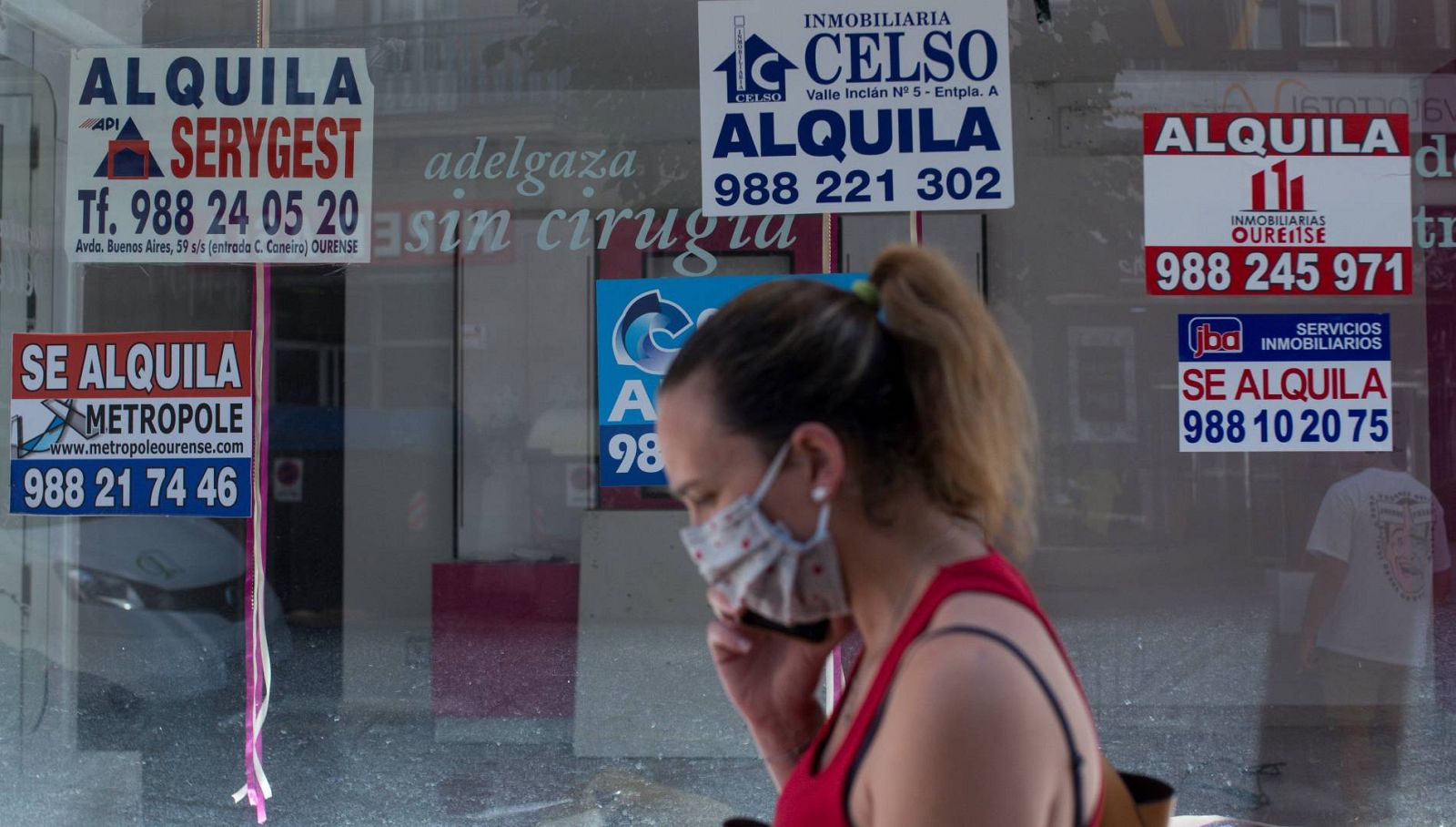 Una mujer con mascarilla pasa ante un comercio cerrado con varios carteles que anuncian su alquiler.