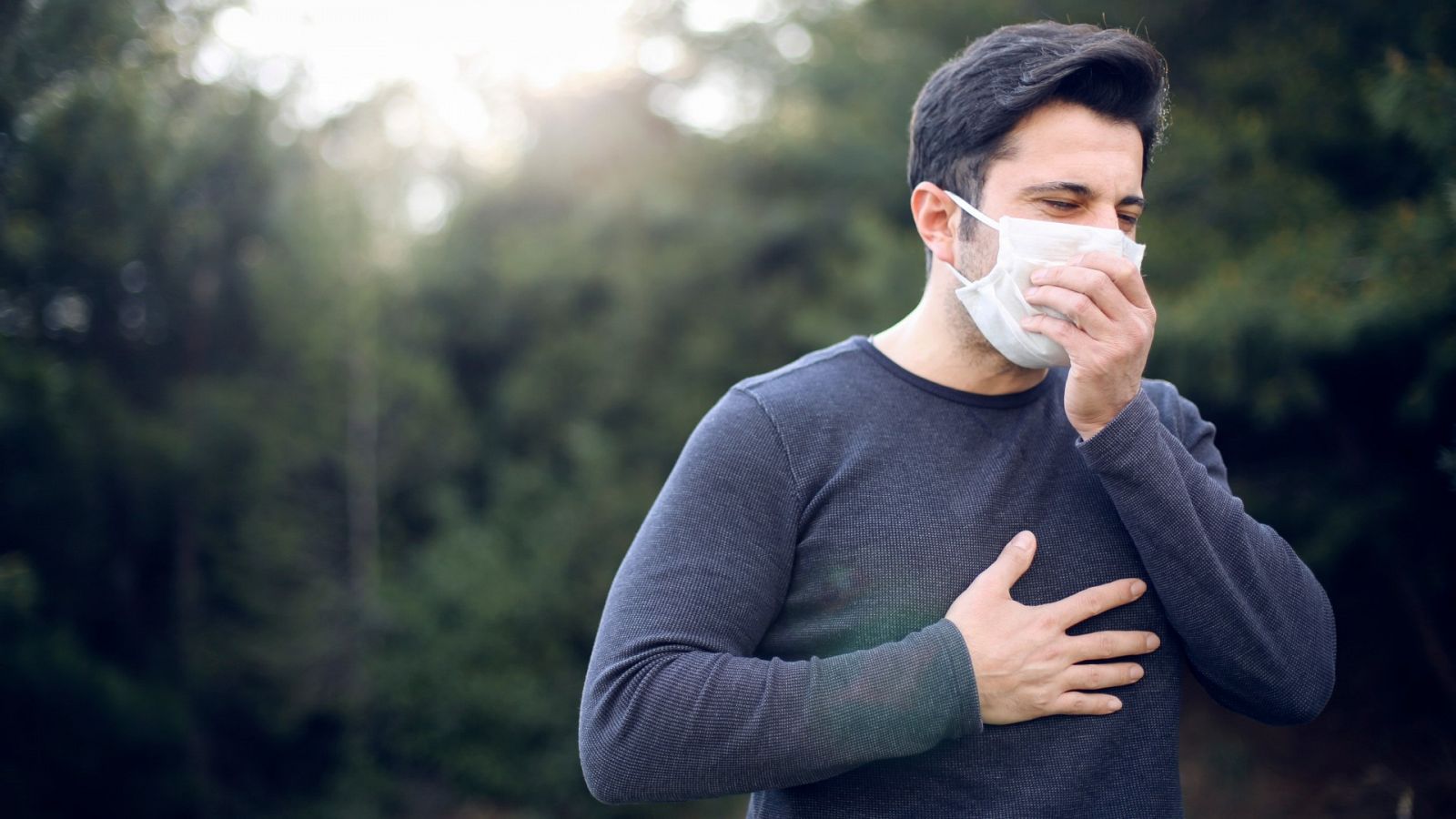 Un hombre con mascarilla tose mientras se lleva la mano al pecho