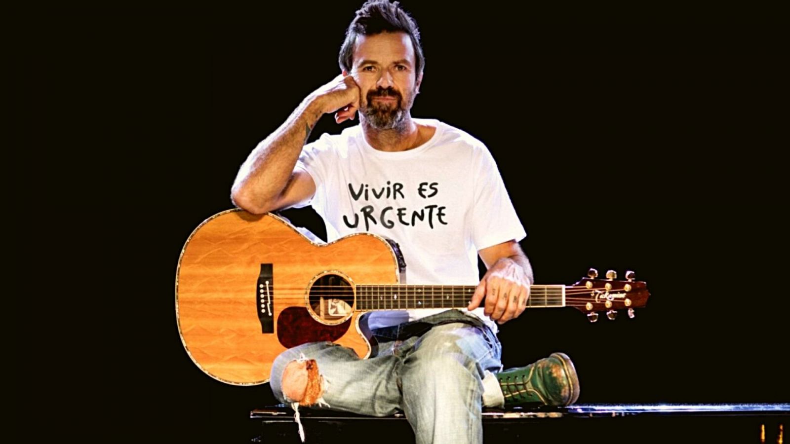 Pau Donés con una camiseta blanca con el mensaje 'Vivir es urgente'. Está sentado sobre un piano de cola con una guitarra acústica entre las manos