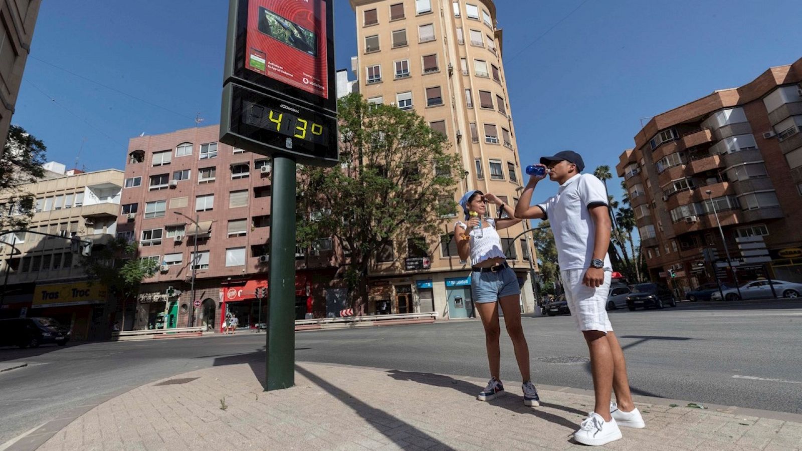 Dos jóvenes beben agua junto a un termómetro que marca 43 grados, en Murcia.