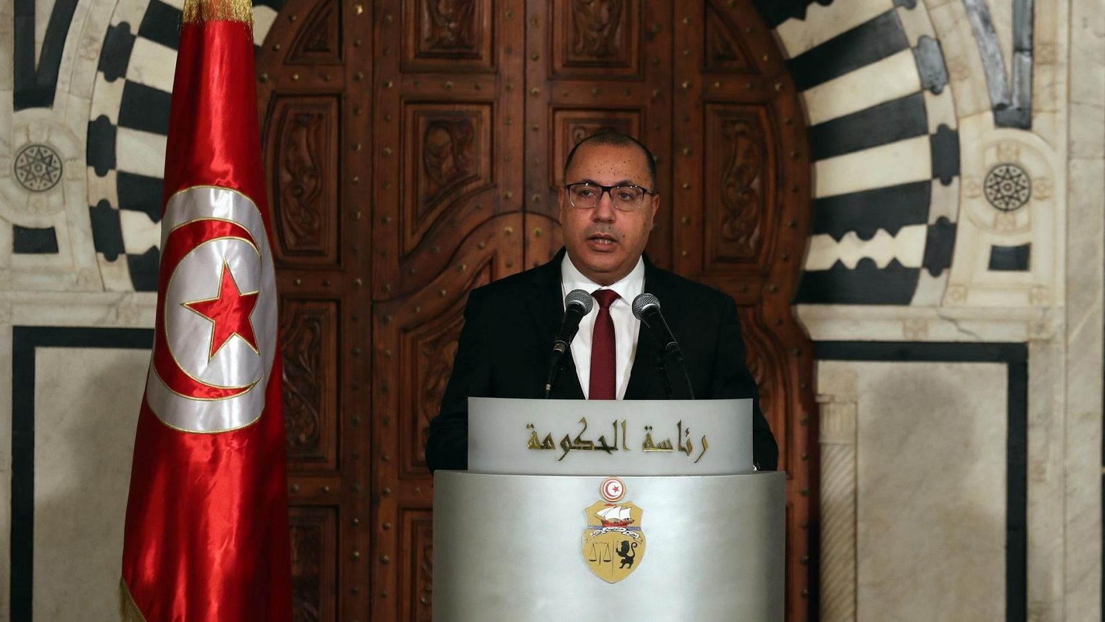 El primer ministro tunecino, Hichem Mechichi habla desde un atril con la bandera del país detrás.