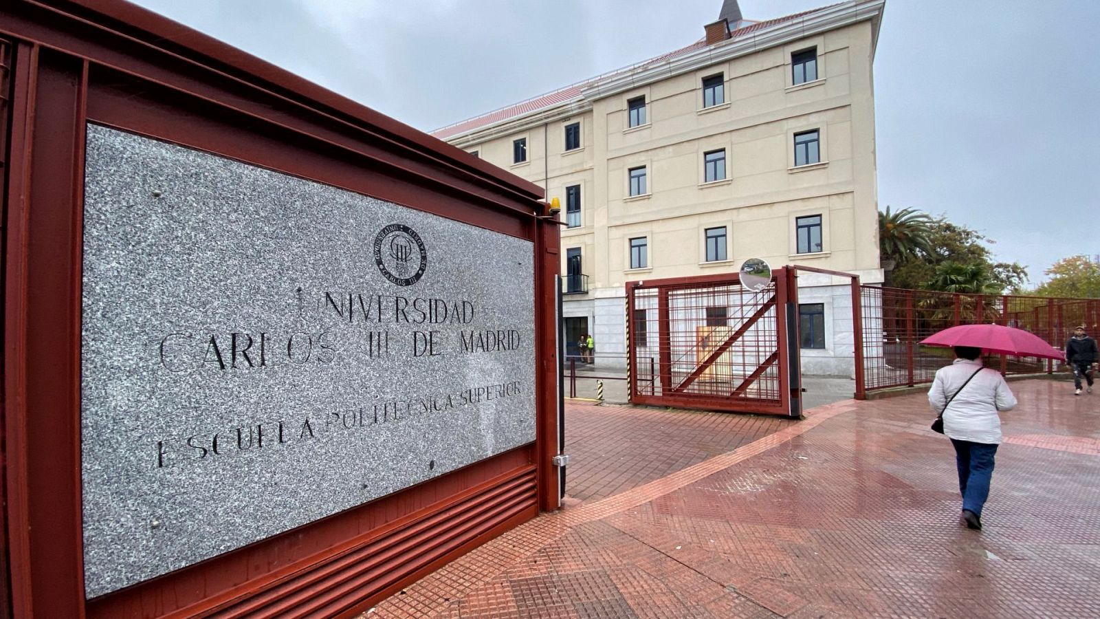 Una mujer con paraguas pasa junto a la placa y la entrada a la Escuela Politécnica Superior del campus de Leganés de la Universidad Carlos III