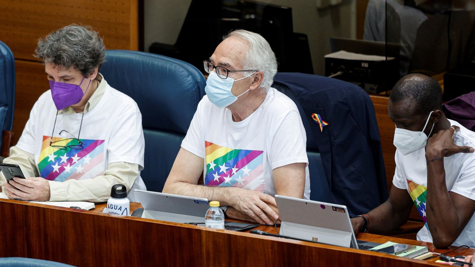 Los diputados de Unidas Podemos Beatriz Gimeno, Agustín Moreno (c) y Serigne Mbaye Diouf con camisetas con las estrellas de la bandera de la Comunidad de Madrid sobre el fondo multicolor de la bandera LGTBI