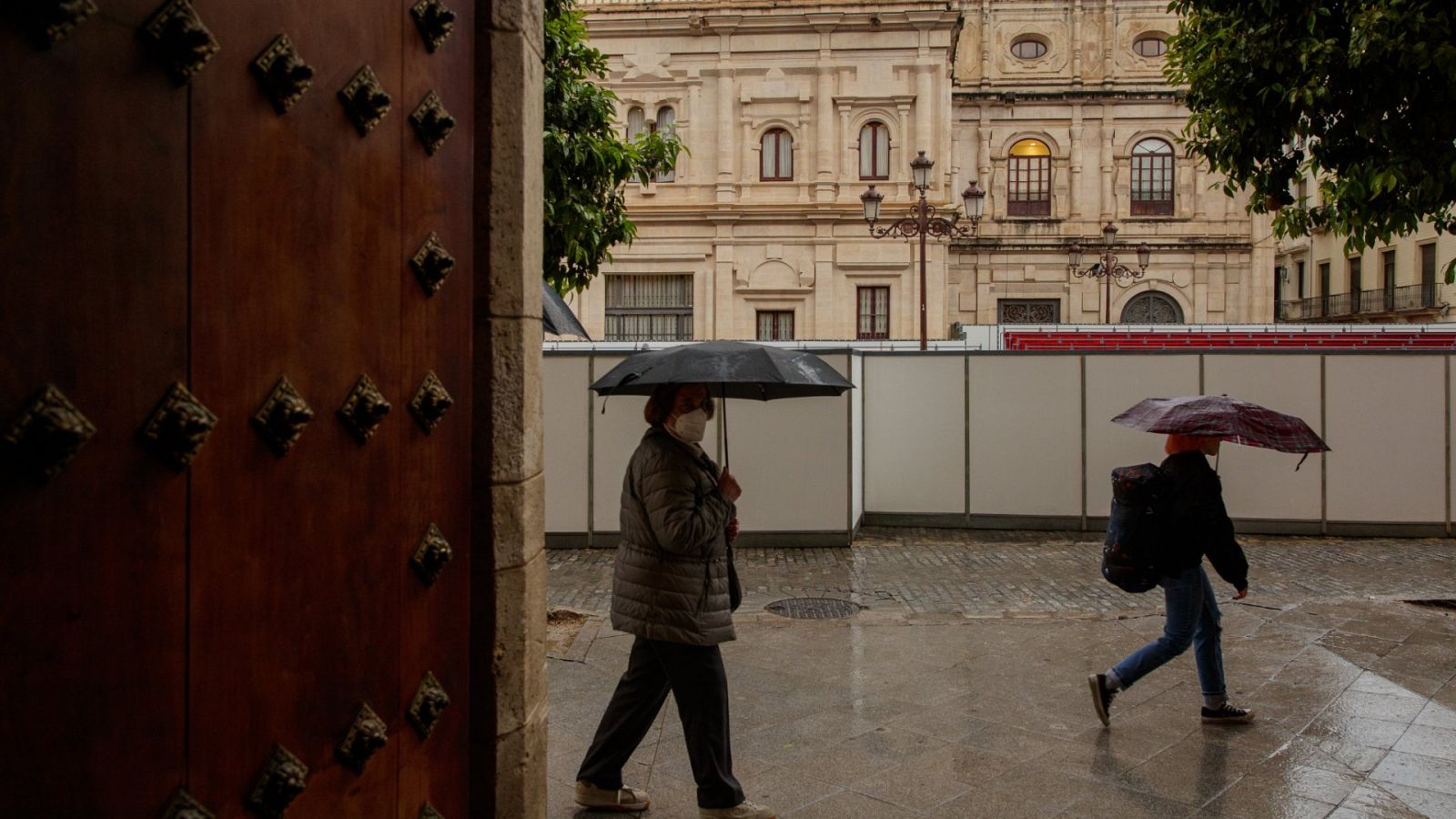 Dos mujeres con paraguas pasan junto a los palcos de Semana Santa instalados ante el Ayuntamiento de Sevilla