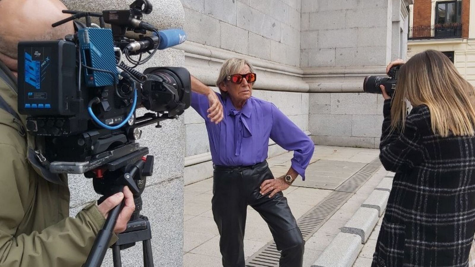 Un anciano con camisa morada posa mientras su nieta le fotografía y la cámara de TVE graba la escena.