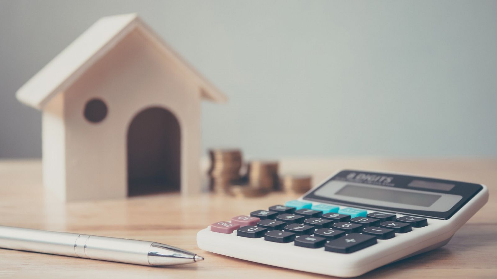Una calculadora con una pila de monedas, una casa de madera y una pluma sobre una mesa