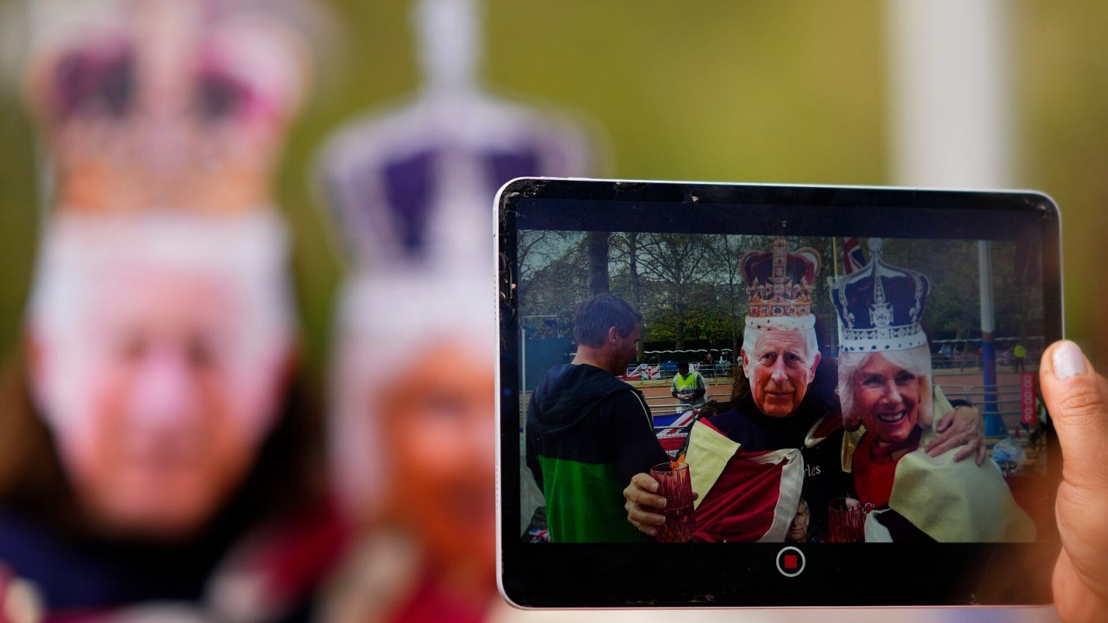 Dos personas posan con las máscaras del rey Carlos III y la reina Camila para una foto con una tablet