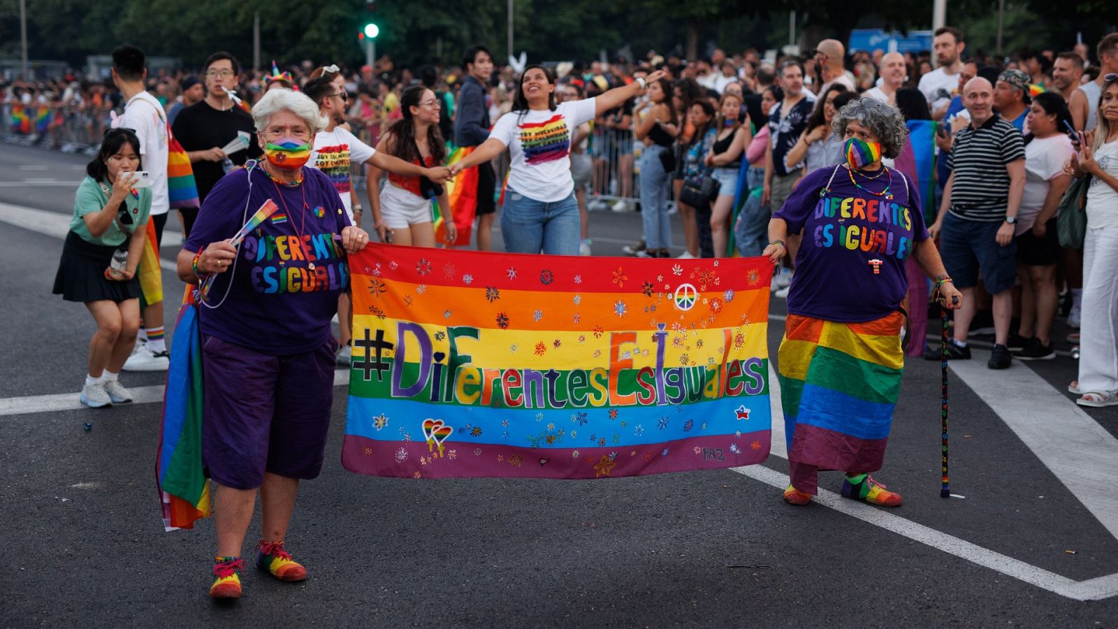 Dos mujeres de mediana edad y vestidas con los colores del Orgullo sujetan una bandera con el lema "Diferentes es Iguales"