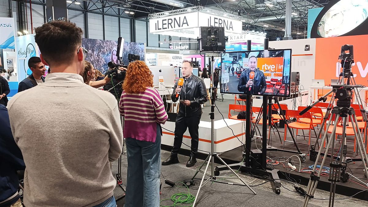 Entrevista en directo o grabada en un stand de ILERNA, con un periodista usando un micrófono naranja y una pantalla mostrando el logo de RTVE.