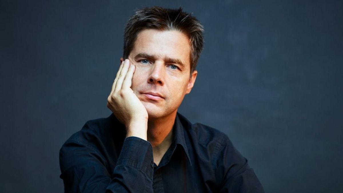 El director de orquesta Christoph König posa con la mano en la cara sobre un fondo oscuro.