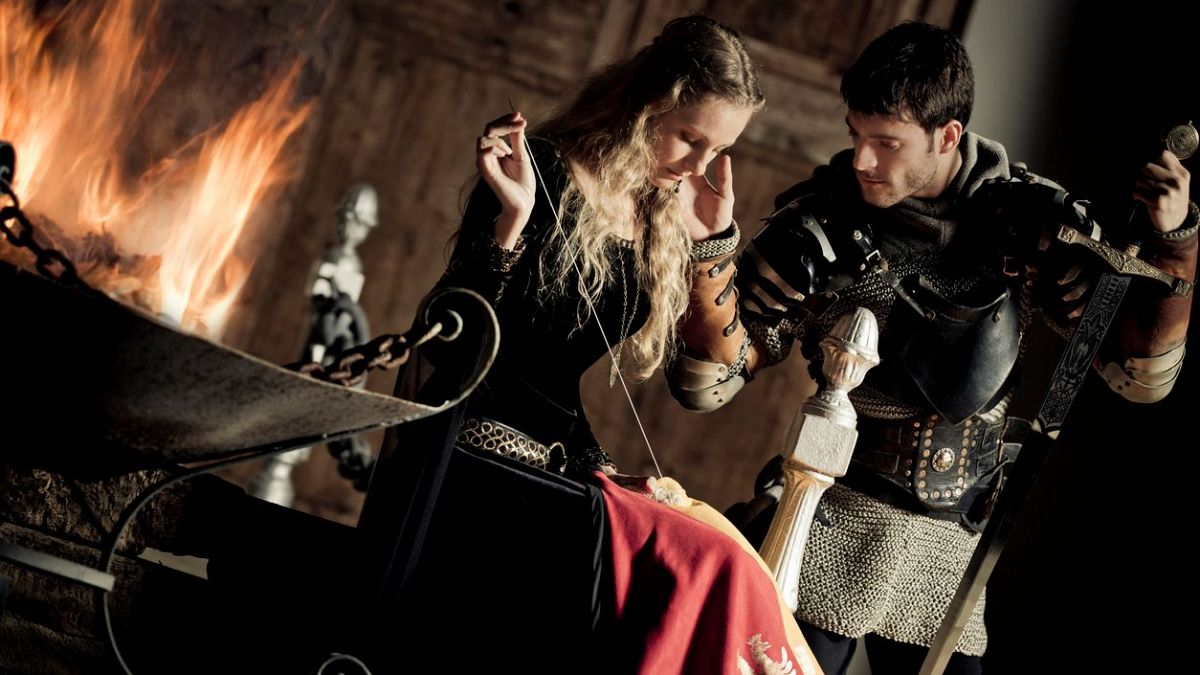 Figurines medievales: mujer cosiendo/tejiendo y hombre con espada y armadura, en un interior con chimenea.  Preparación para una escena.