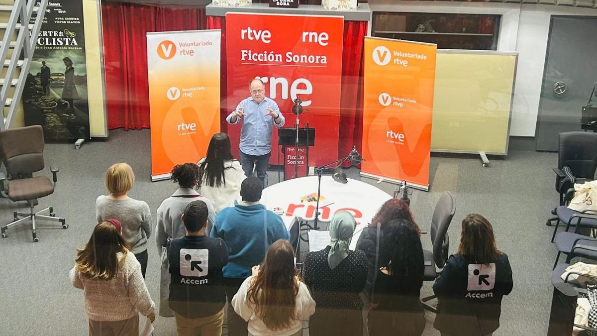 Grabación de ficción sonora en estudio. Grupo de personas alrededor de una mesa con micrófonos, dirigidos por un hombre.  Carteles de RTVE, RNE y 'Voluntariado' presentes.