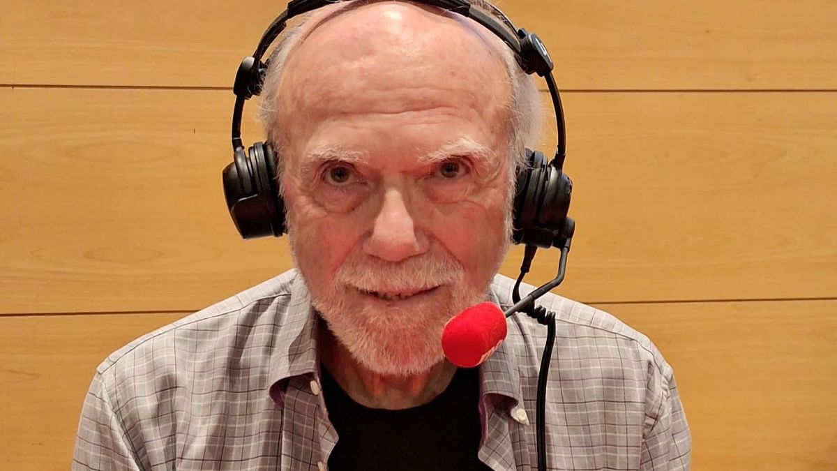 Hombre mayor con auriculares y micrófono, cabello y barba blancos, camisa a cuadros, grabando audio o entrevista. Fondo de madera clara.