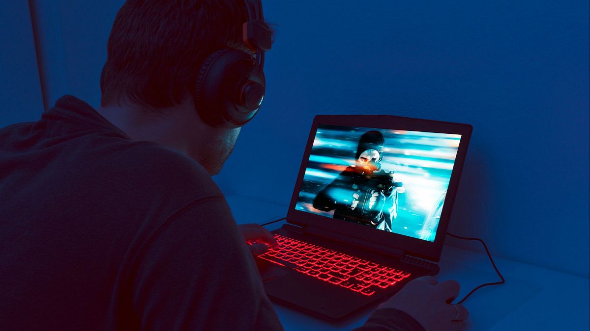 Hombre con auriculares jugando a un videojuego en su portátil.  Pantalla con escena de acción y efectos luminosos; teclado con retroiluminación roja en habitación oscura.