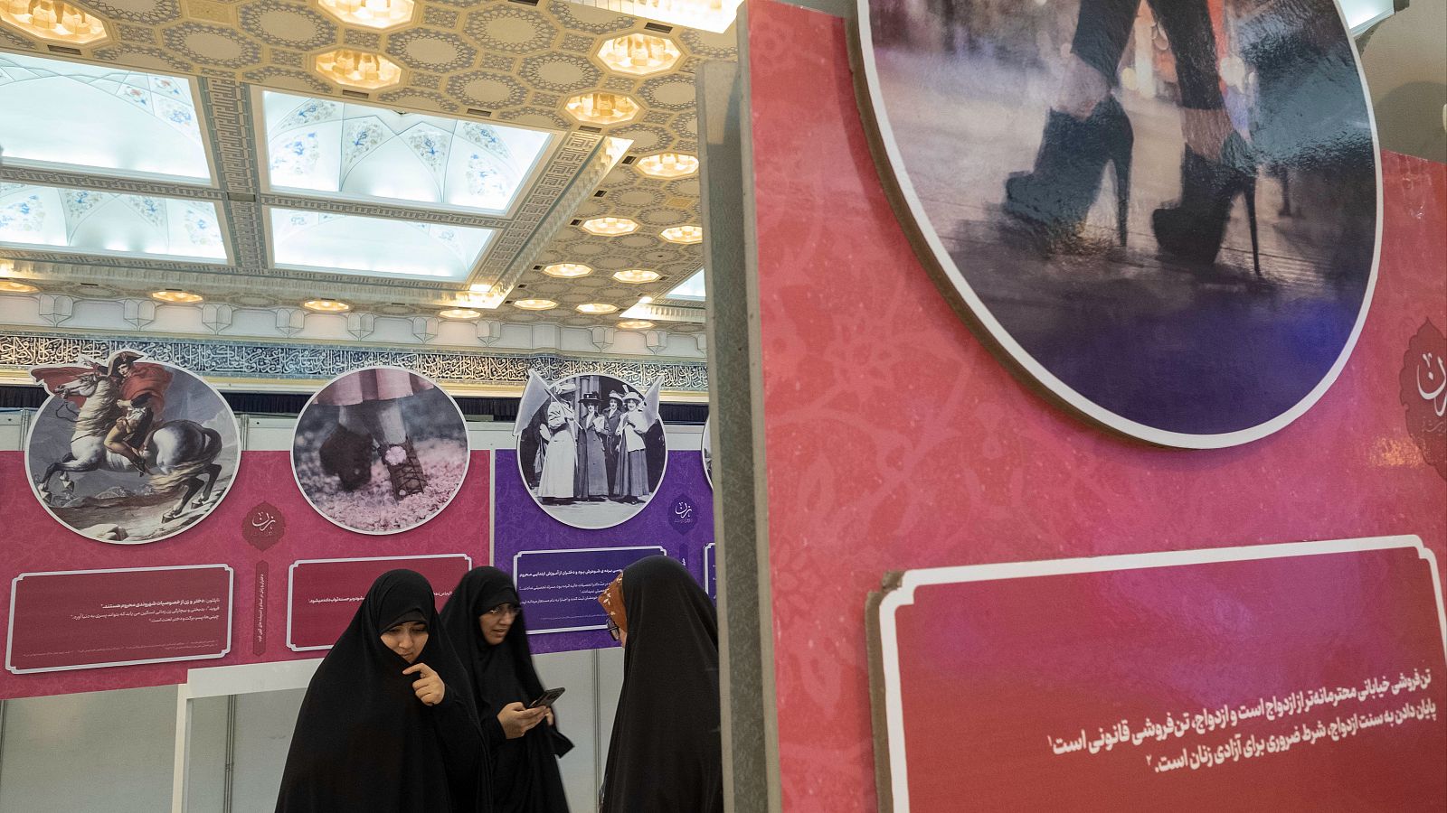Mujeres iraníes con velo se encuentran junto a un cartel (derecha) con la imagen de las piernas de una mujer iraní y una escritura persa que dice: "La prostitución callejera es más respetable que el matrimonio, y el matrimonio es prostitución legal"