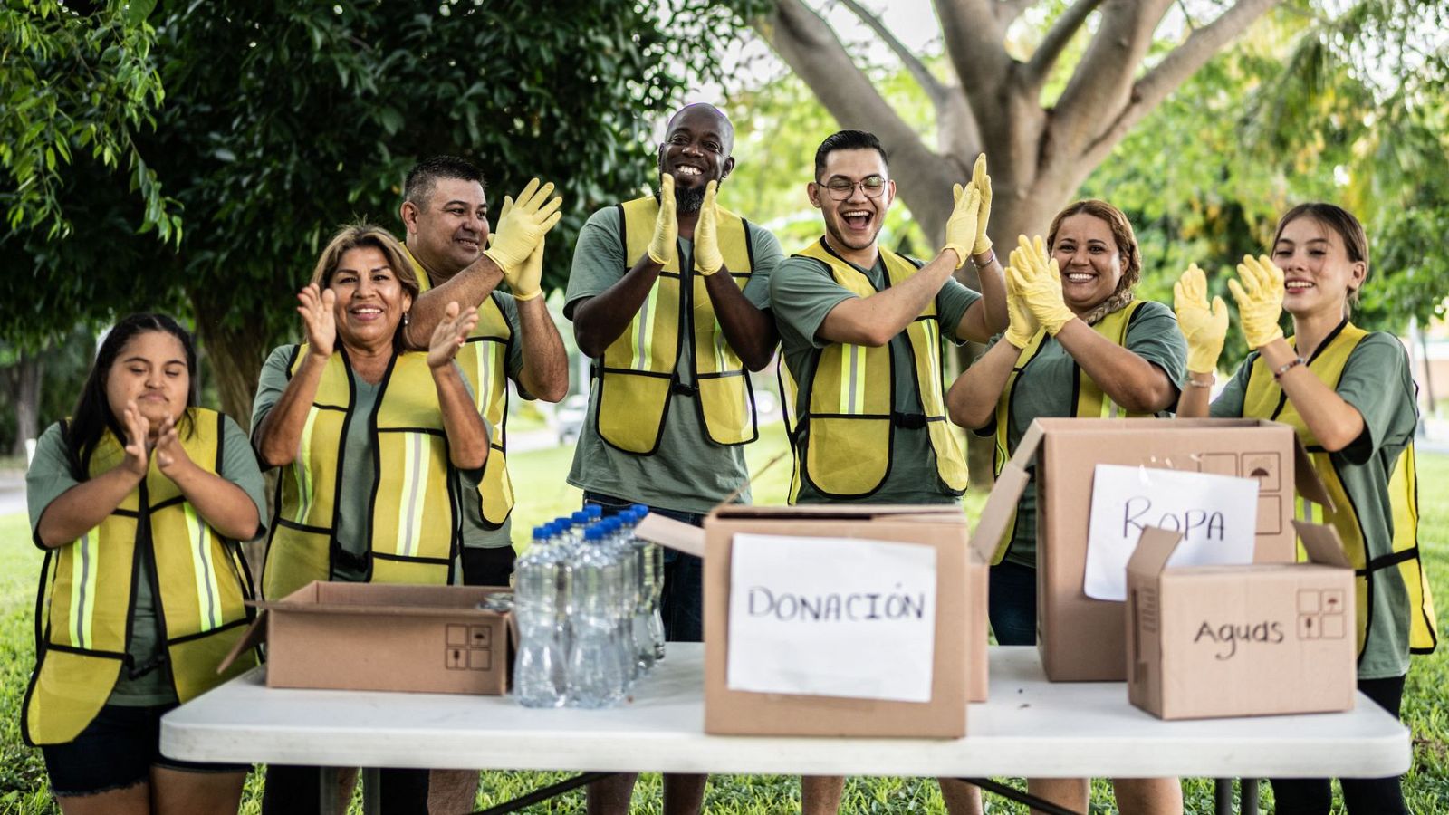 Un grupo de personas con chalecos amarillos aplaude junto a unas cajas de cartón con etiquetas de ropa, agua y donaciones.