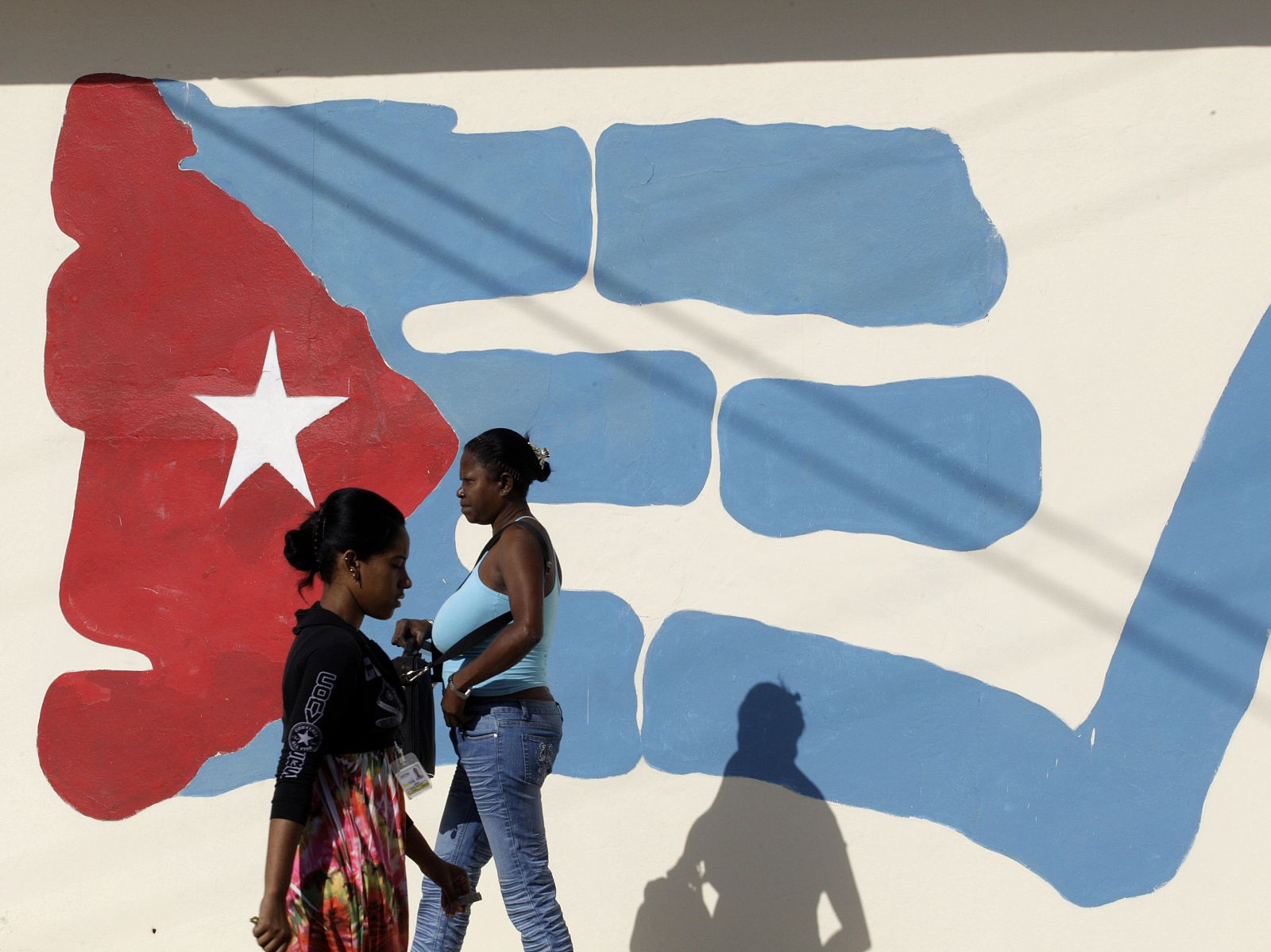 Dos mujeres pasean junto a una pared con un graffiti de la bandera cubana.