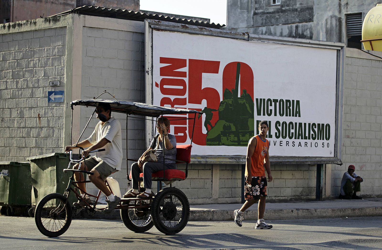 Un hombre lleva a una mujer en su triciclo taxi junto a un cartel que celebra el 50 aniversacio del incidente de Bahía de Cochinos.