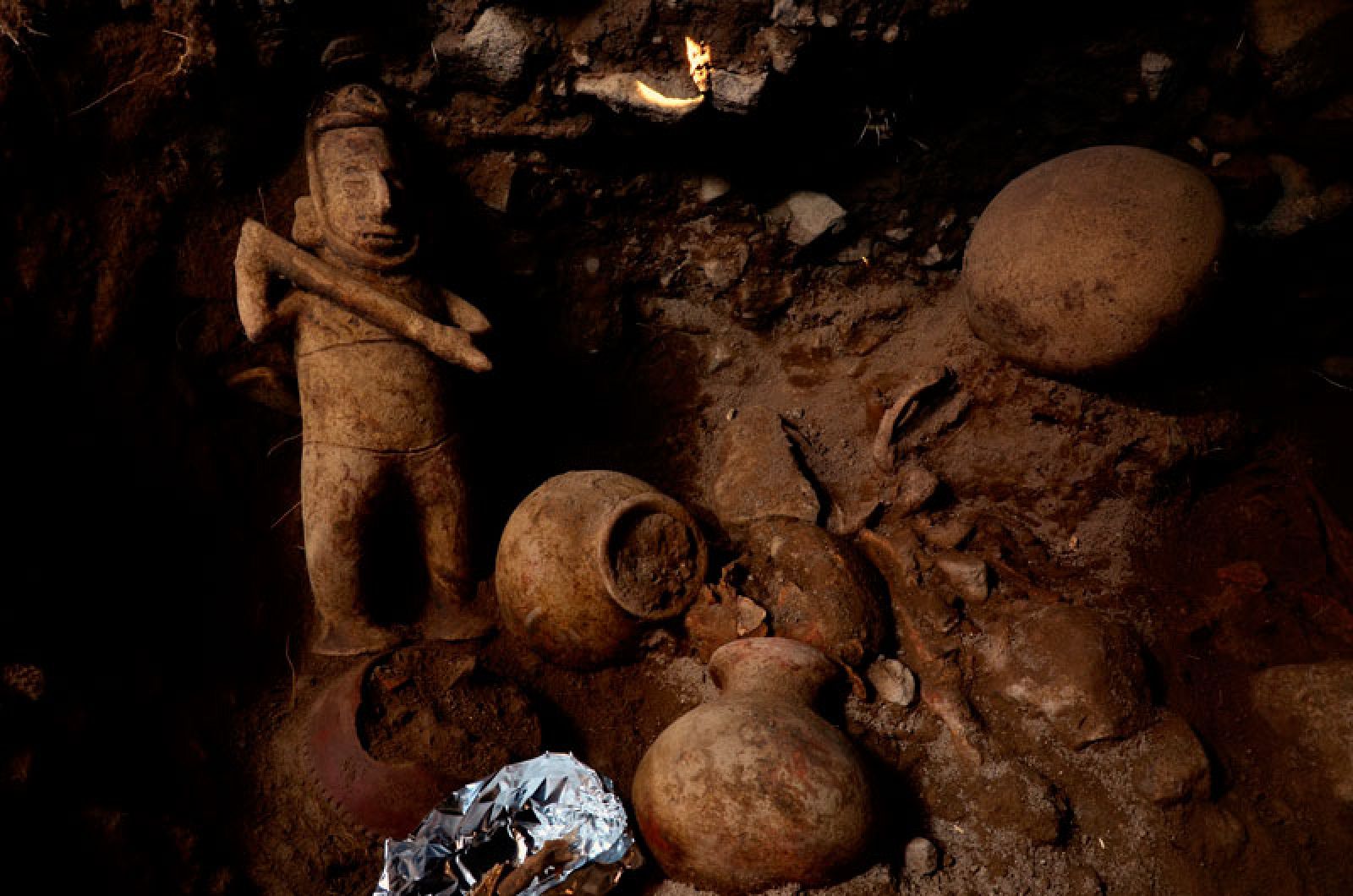 Escultura de un chamán de rostro alargado y arma en mano, junto a vasijas halladas.
