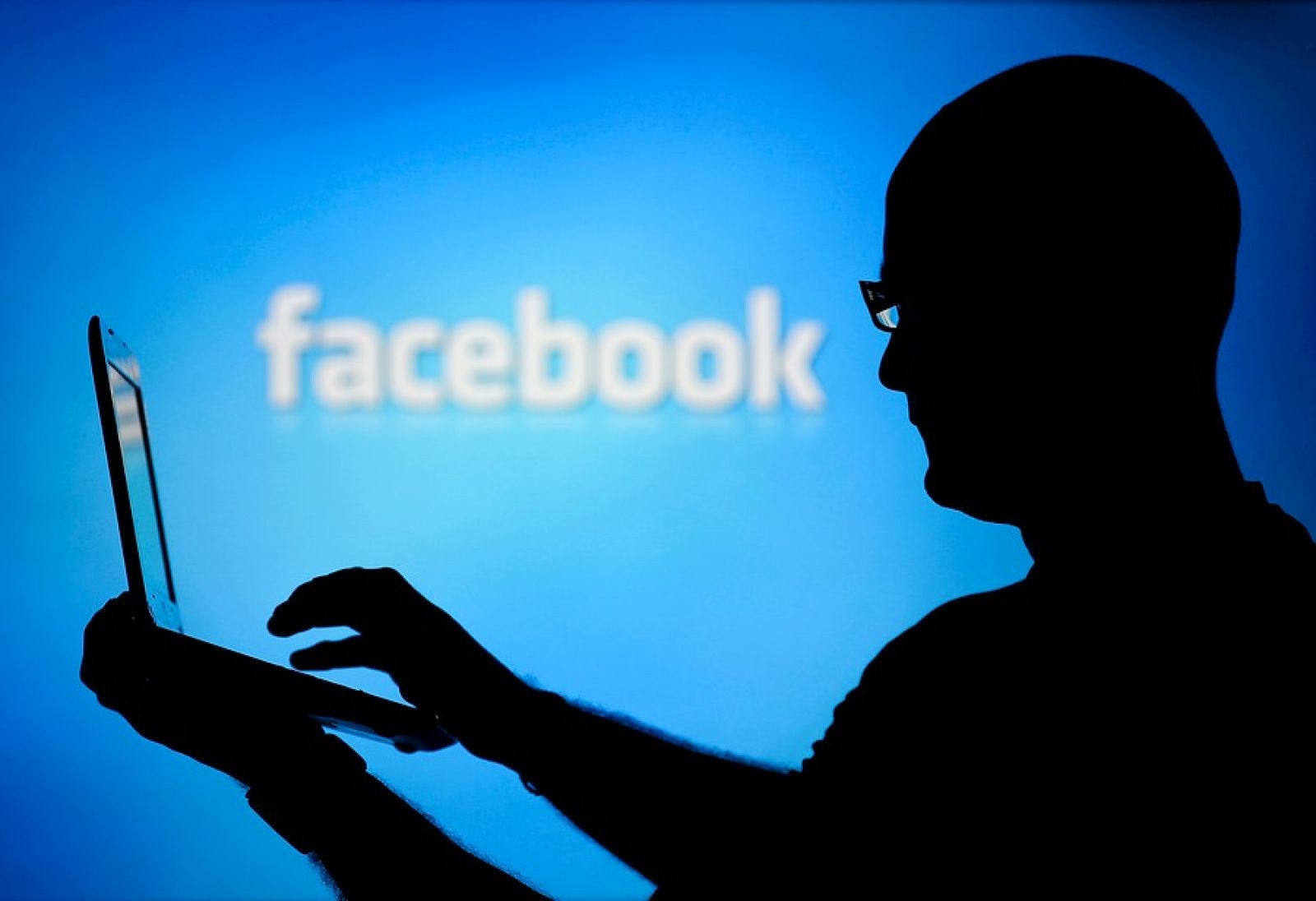 Hombre con un ordenador y el logo de Facebook de fondo.