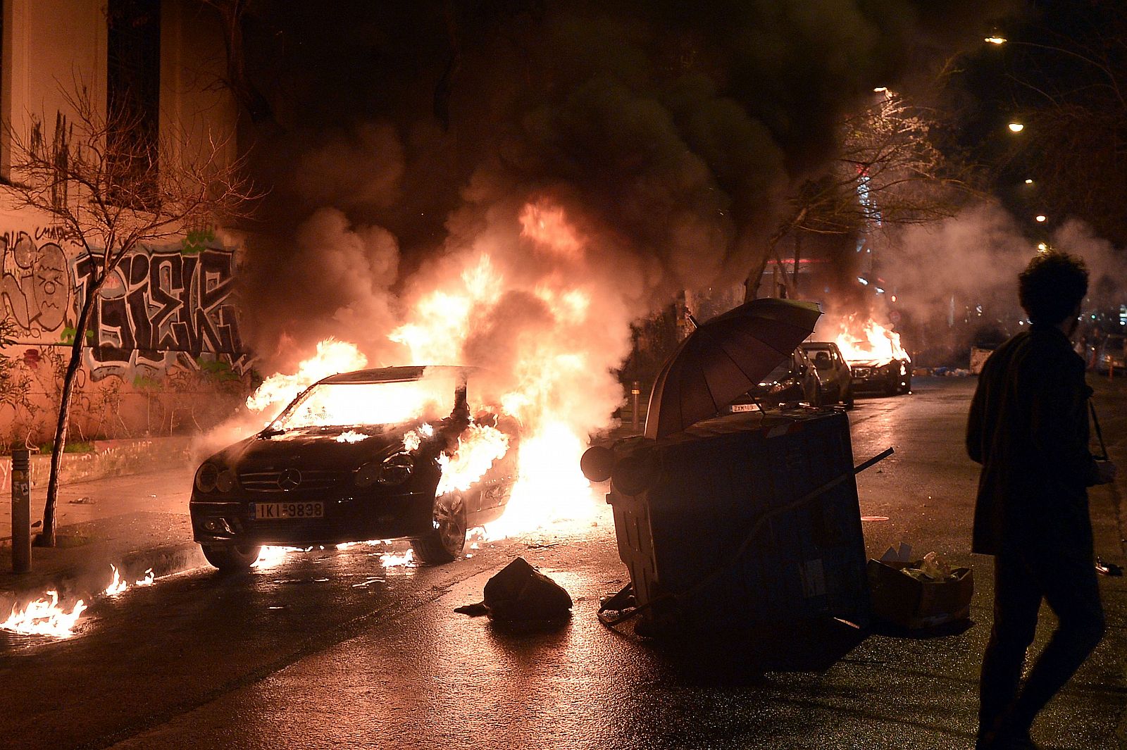 Un hombre en capuchado se aleja de un coche ardiendo en Atenas después de que otro joven lo quemase al término de una manifestación.
