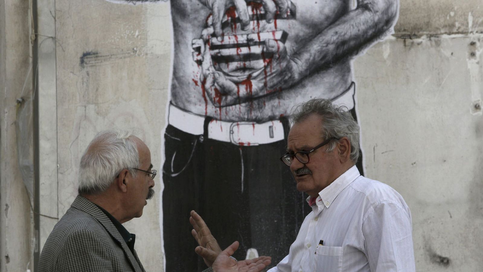 Dos hombres conversan en Atenas, delante de un graffiti que muestra un hombre que sujeta un símbolo del euro ensangrentado
