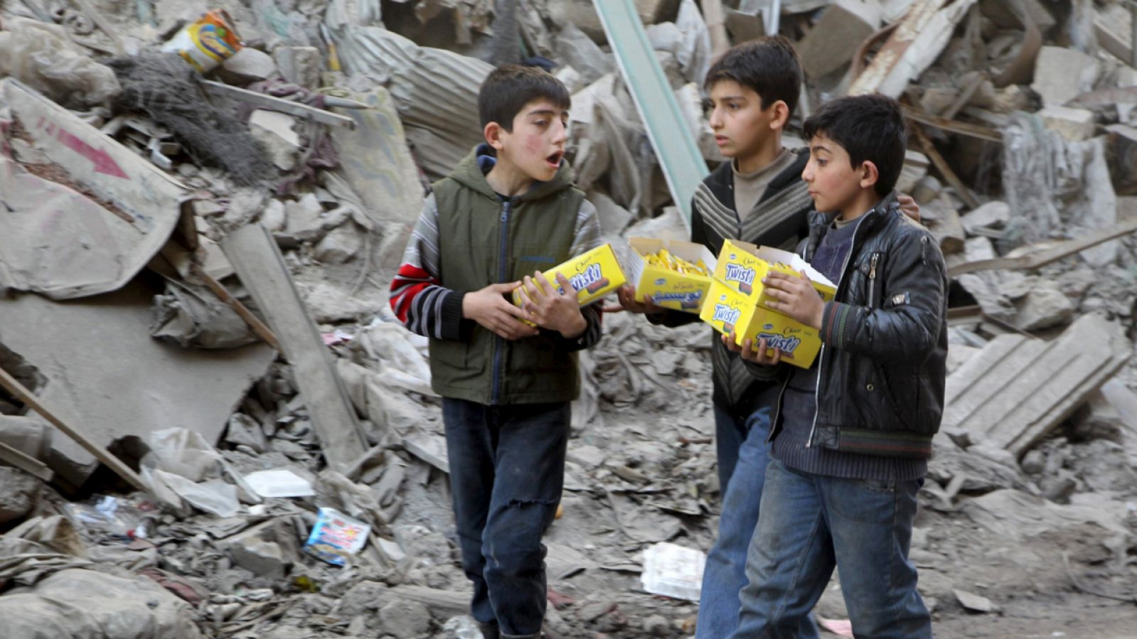 Niños sirios llevan cajas de galletas junto a las ruinas de un edificio en Alepo