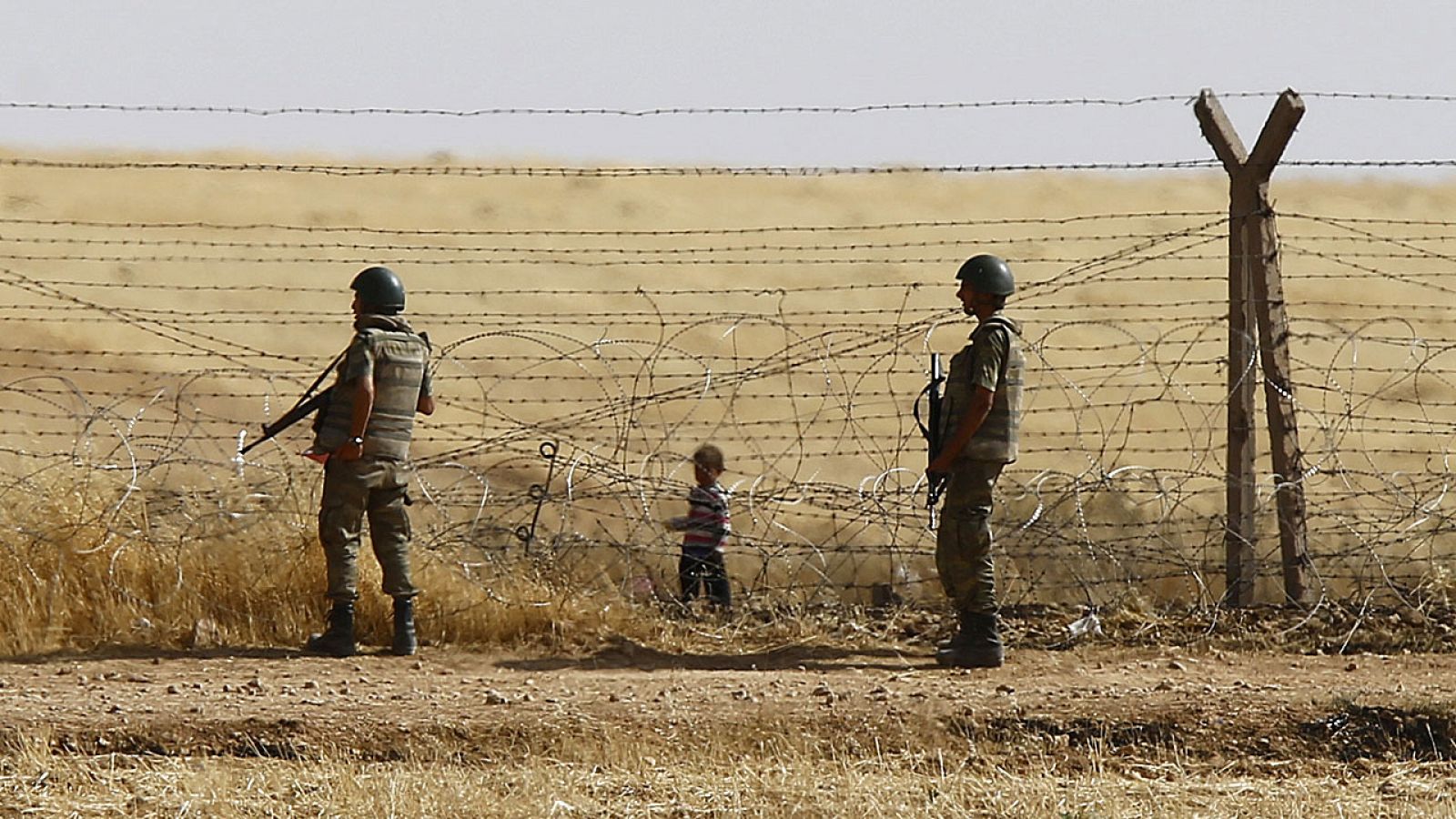 Soldados turcos hacen guardia mientras un niño refugiado sirio camina cerca de la valla fronteriza.