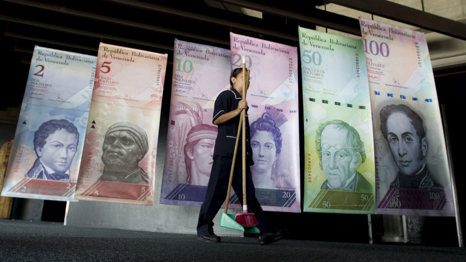 Una mujer pasa con utensilios de limpieza frente a afiches de billetes venezolanos en la sede del Banco Central de Venezuala en Caracas