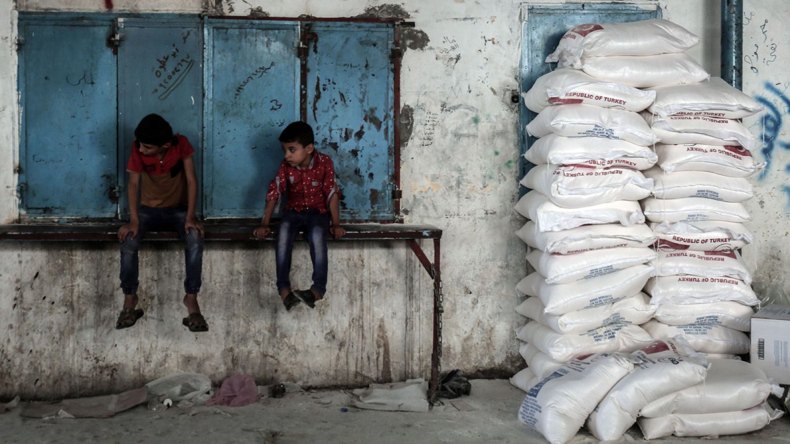 Dos niños gazatíes esperan sentados al lado de sacos de comida provenientes de la ayuda humanitaria de Agencia de Naciones Unidas para los Refugiados de Palestina en Oriente Próximo.