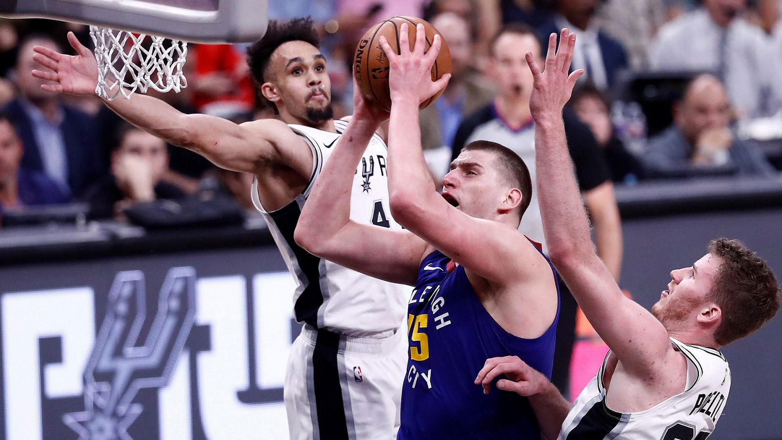 El jugador de los Nuggets de Denver, Nikola Jokic de Serbia va a la canasta contra los jugadores de los San Antonio Spurs Derrick White y Jakob Poeltl.