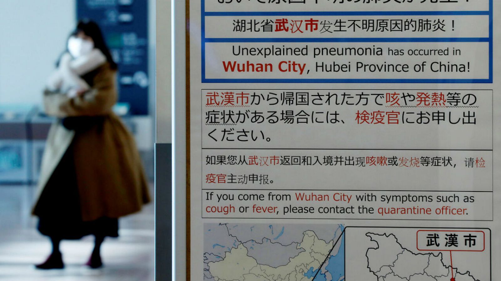Una mujer camina con una máscara, dejando atrás un panel de información en el que se advierte del brote de coronavirus en China, en el aeropuerto Haneda, en Tokio.