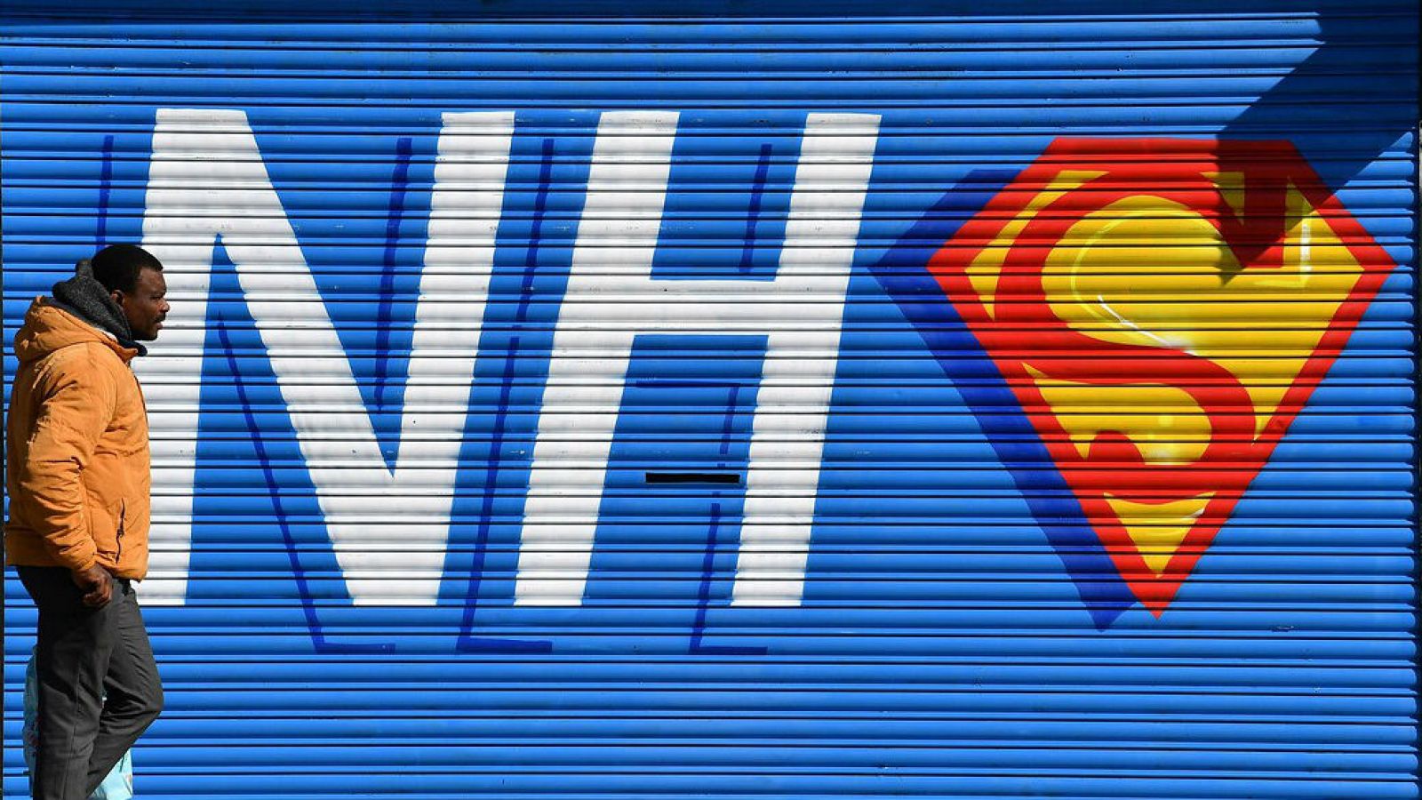 Un peatón pasa junto a un graffiti que representa el logotipo del NHS (Servicio Nacional de Salud) de Gran Bretaña, fusionado con el emblema o insignia del superhéroe ficticio Superman y Superwoman, en la entrada cerrada de un restaurante de pizza ce