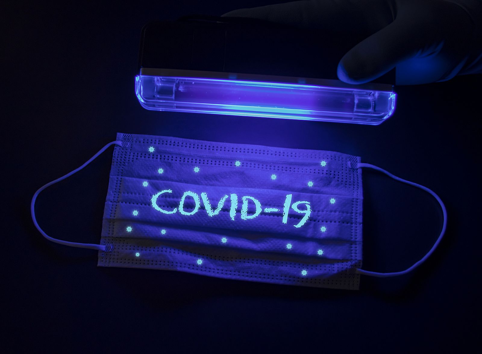 Mascarilla quirúrgica con la inscripción Covid-19 y con unas pequeñas moléculas de coronavirus bajo una lámpara ultravioleta.