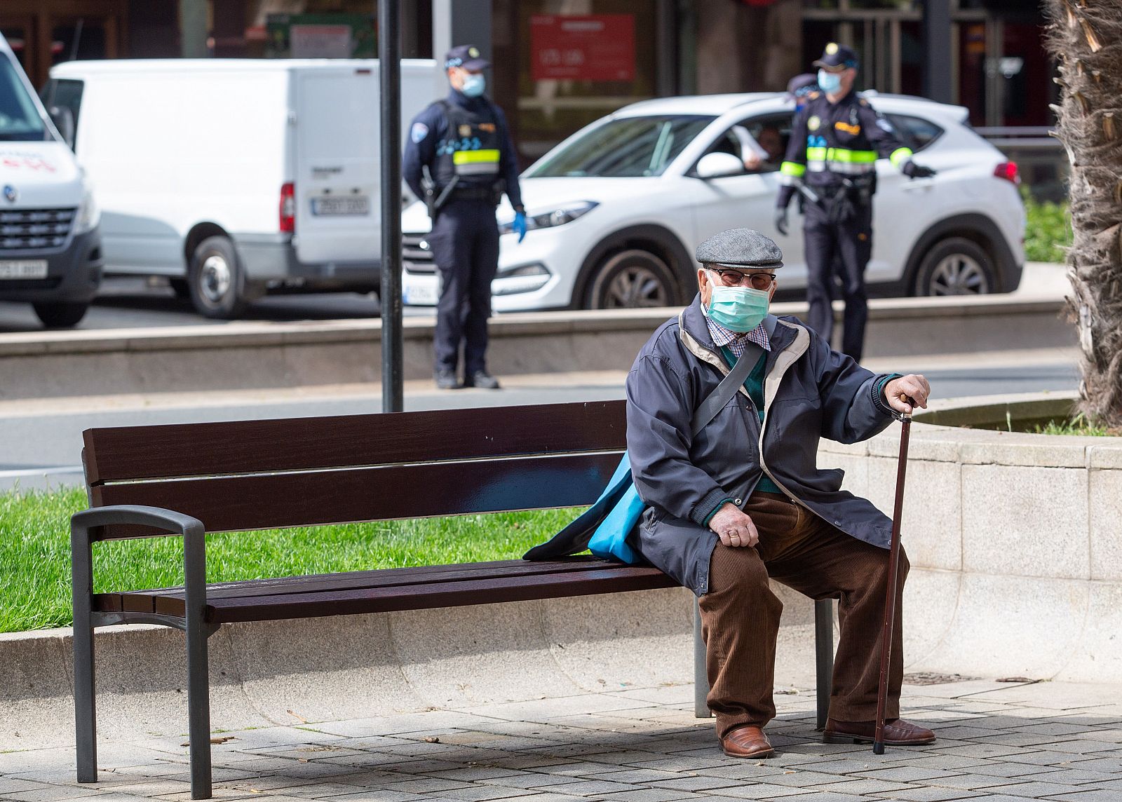 Un hombre con mascarilla permanece sentado en un banco mientras a sus espaldas agentes de la Policía Nacional  trabajan