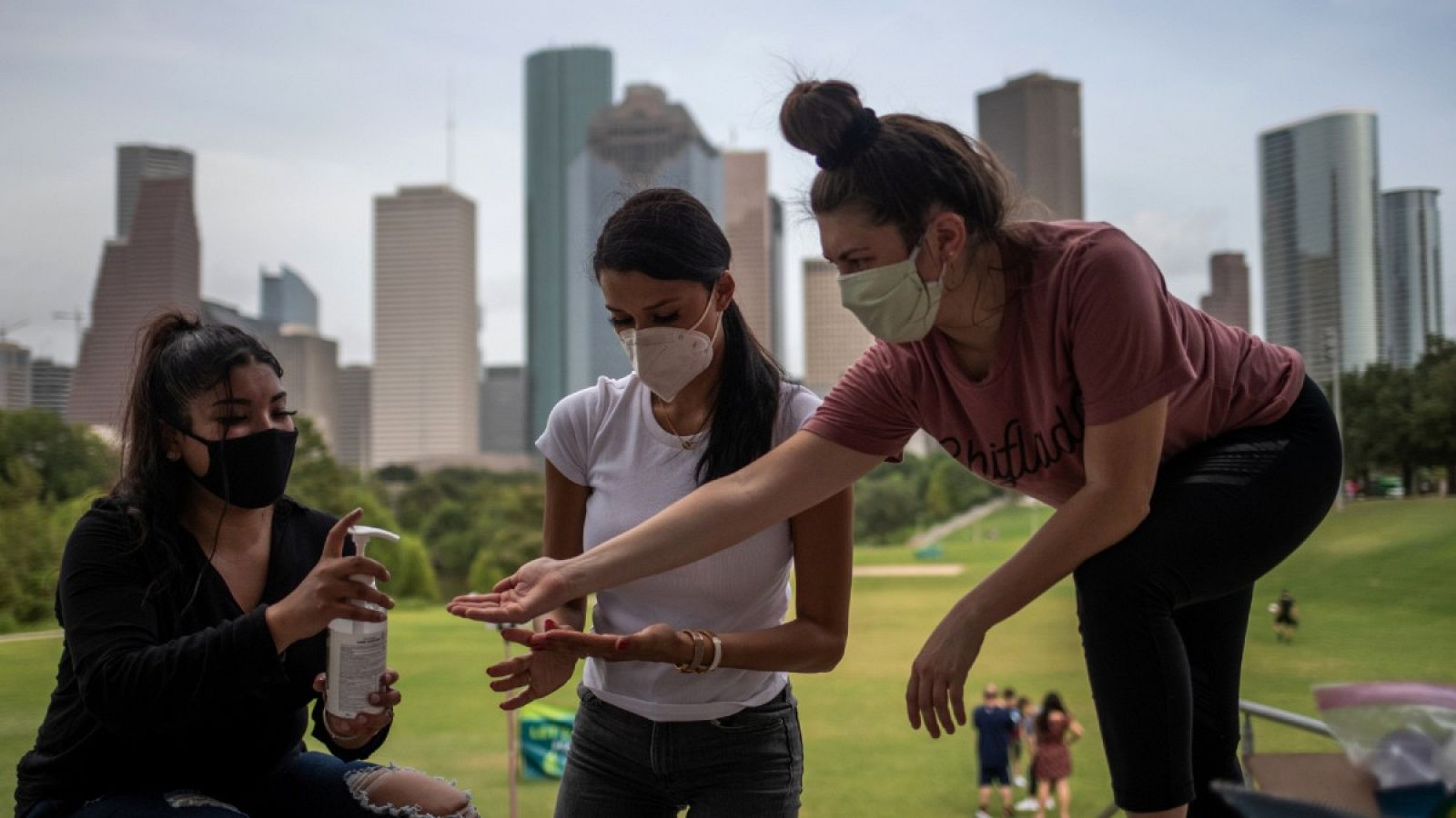 Tres jóvenes con mascarilla comparten desinfectante de manos con el 'skyline' de Houston, Texas, de fondo.