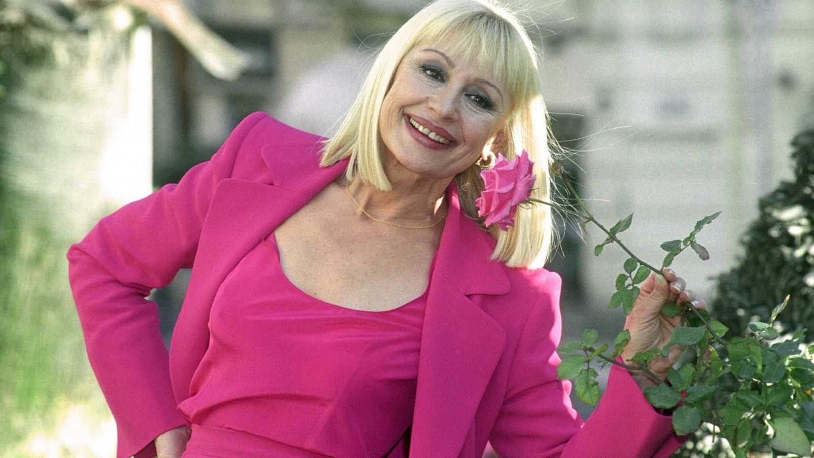 Raffaella Carrà oliendo una flor vestida con un traje rosa fucsia