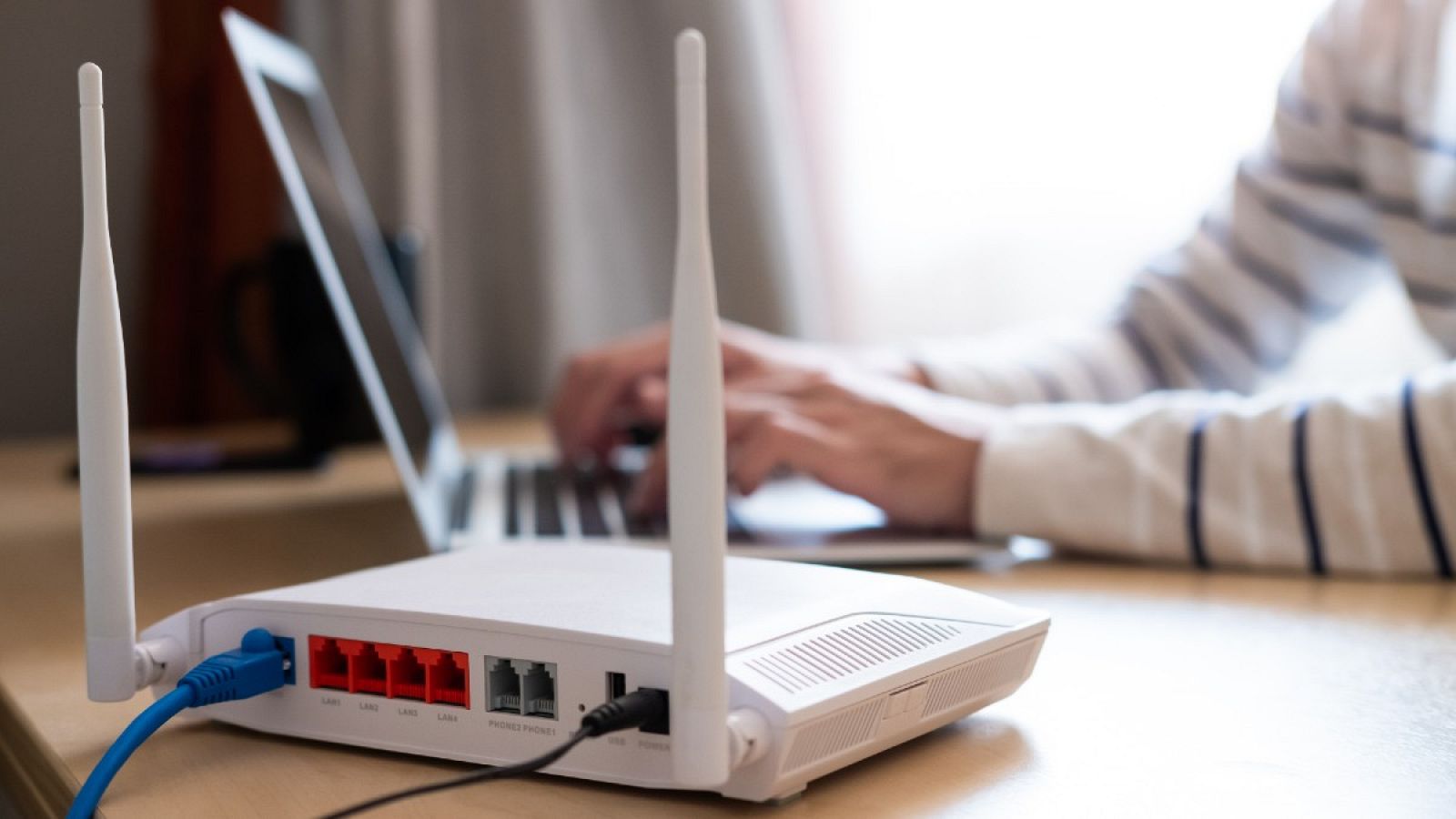 Imagen de un router blanco en primer plano mientras una persona utiliza su ordenador en el fondo
