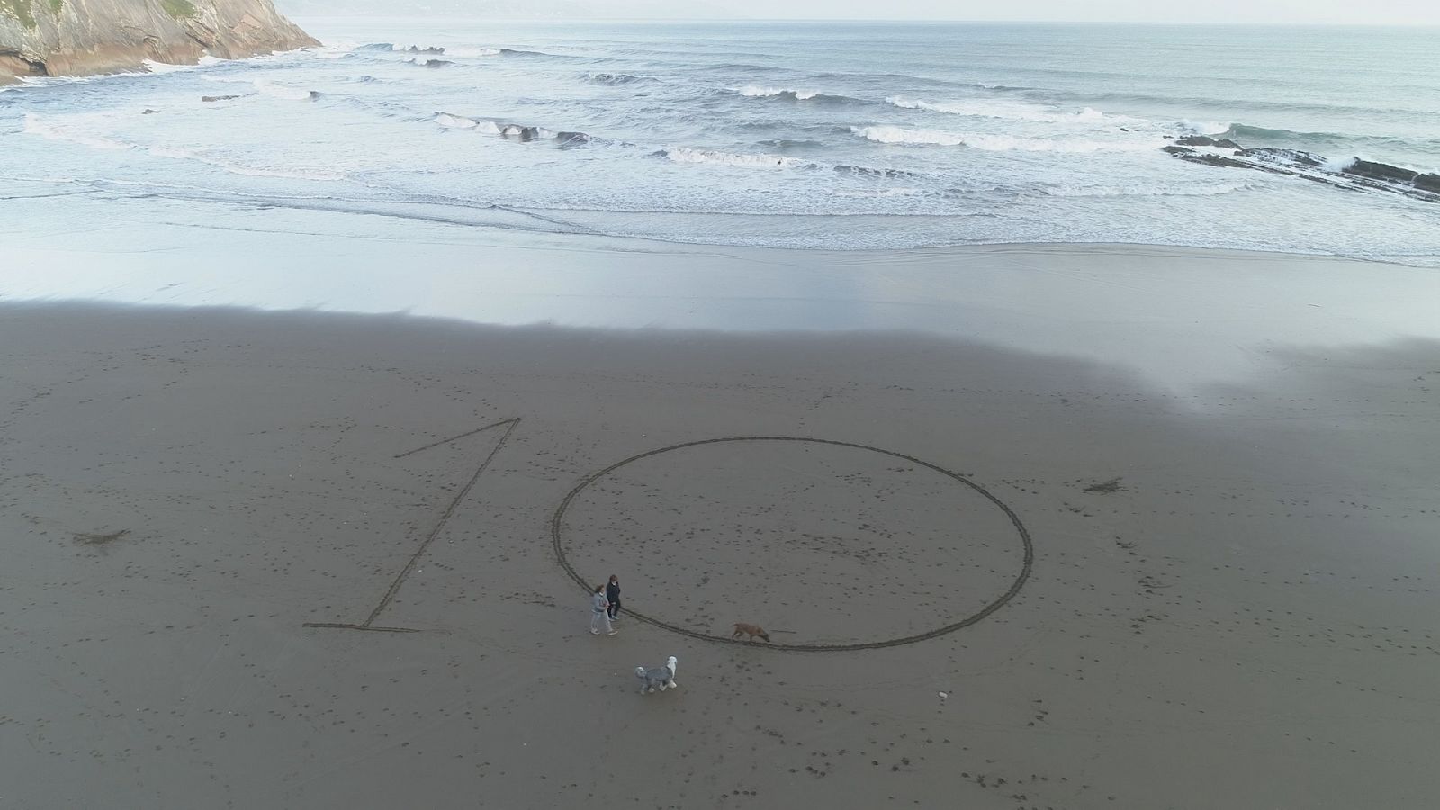 Una pareja y sus perros pasean por la playa donde hay escrito en la arena un diez enorme.
