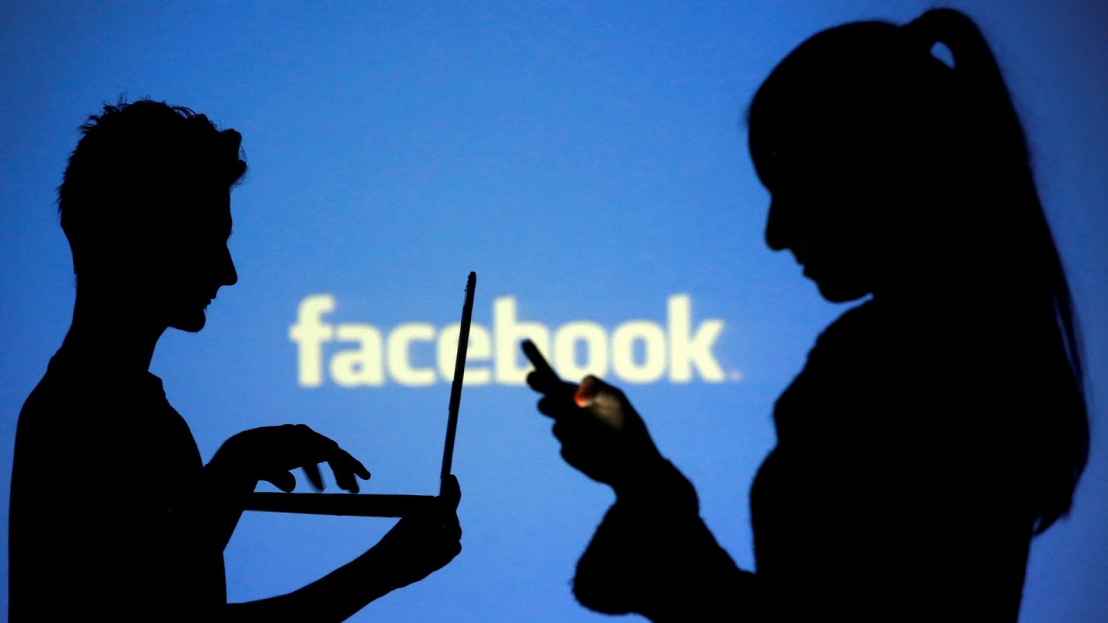 Siluetas de dos personas usando un ordenador y un teléfono móvil frente a una pantalla con el logo de Facebook