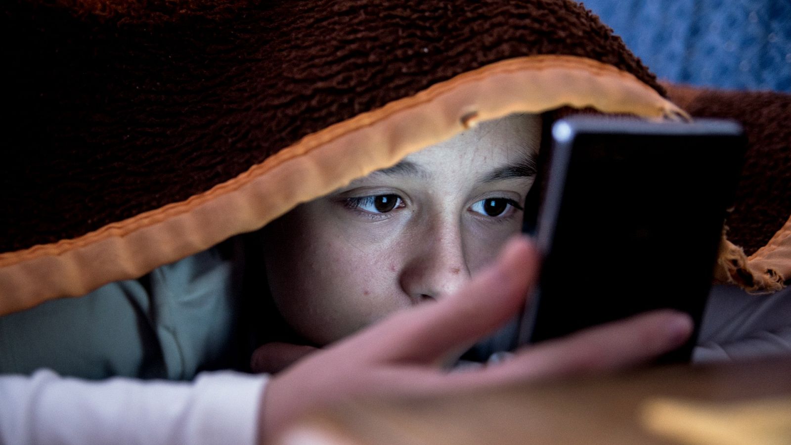 Una adolescente mira la pantalla de su móvil mientras se cubre con la sábana