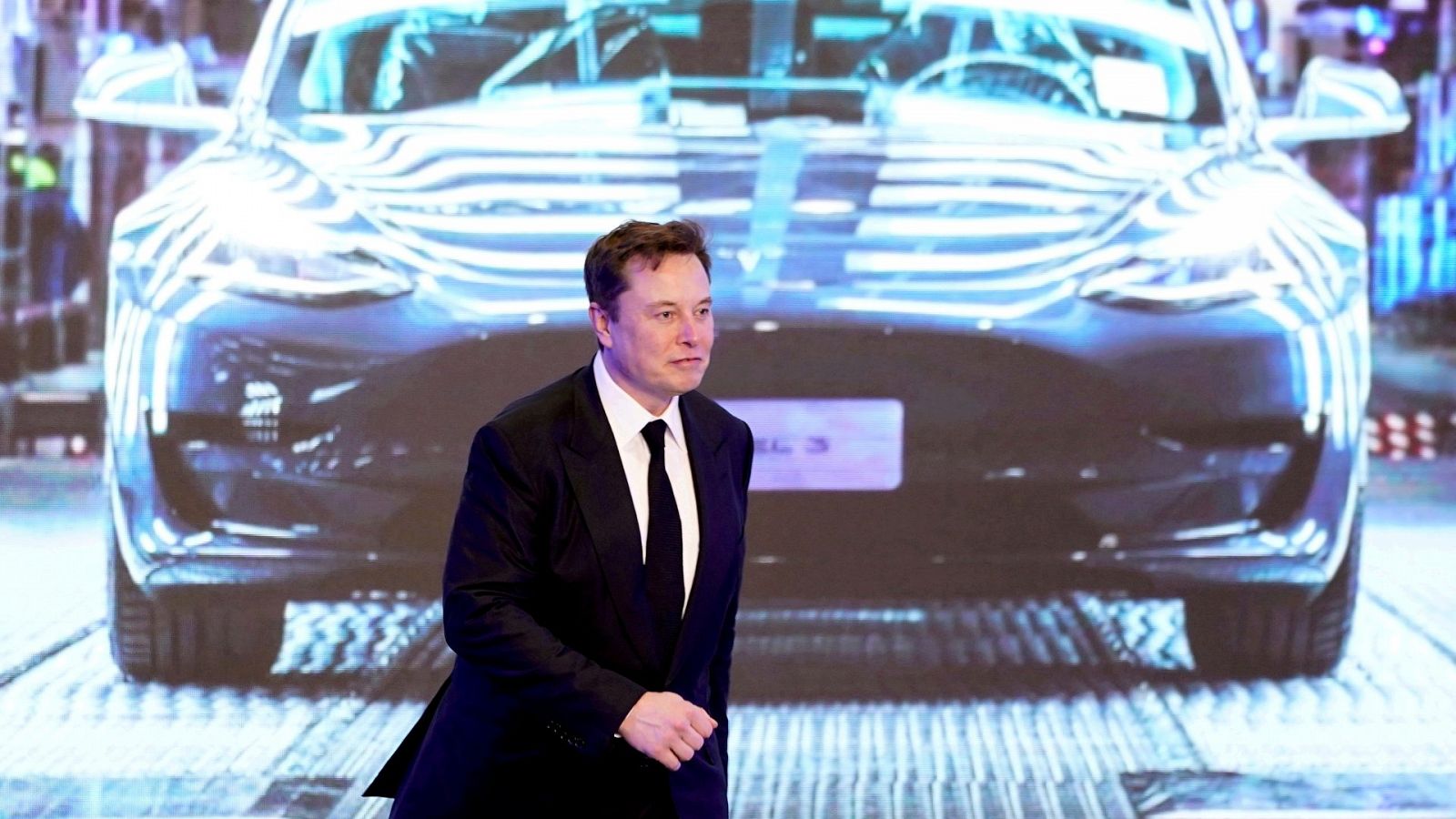 Elon Musk camina junto a una pantalla que muestra una imagen del coche Tesla Model 3