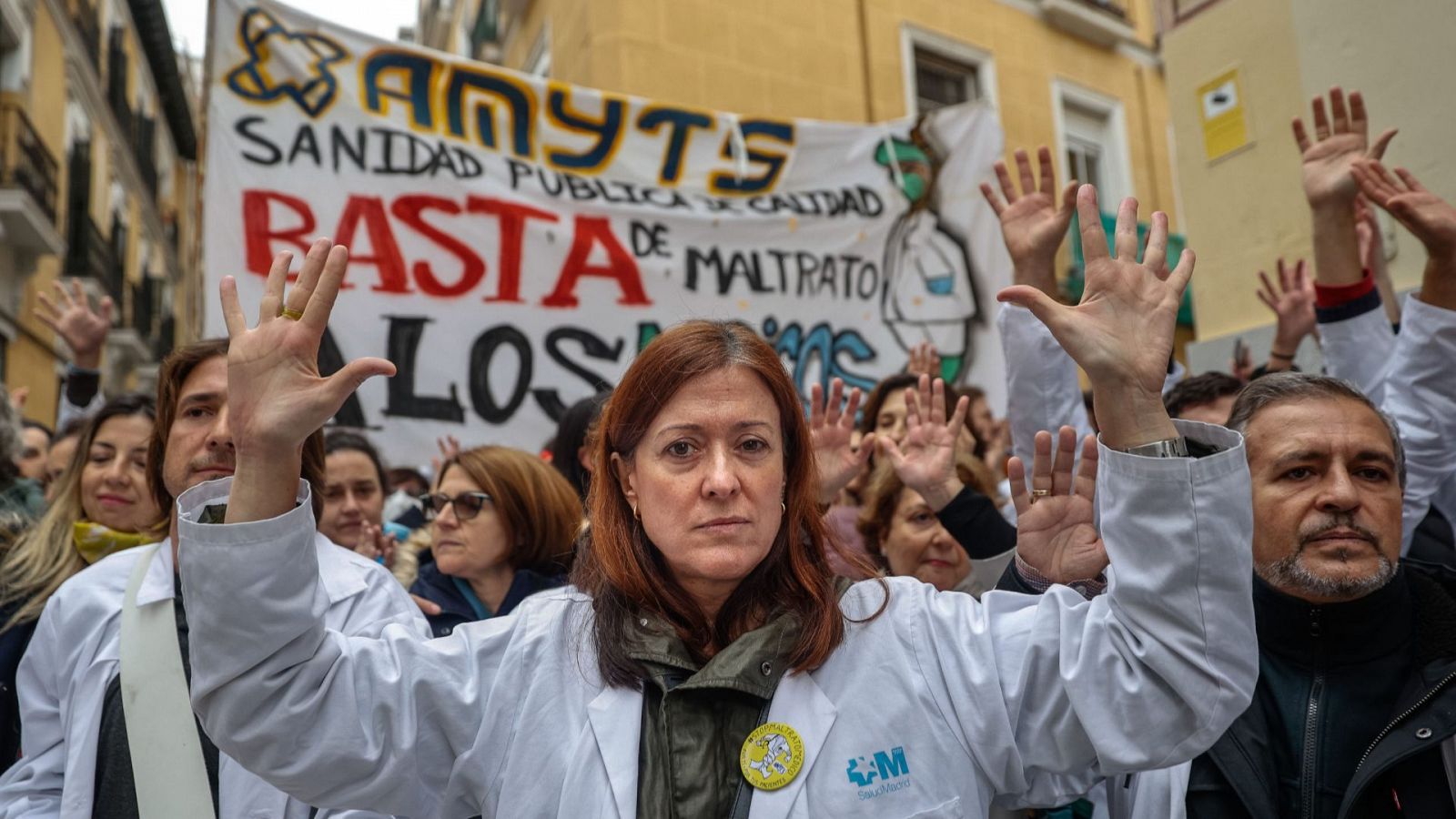 Participantes de la huelga indefinida de médicos de familia y pediatras de la Atención Primaria madrileña. Tienen las manos en alto y atrás una pancarta que dice: "Sanidad pública de calidad. Basta de maltrato"
