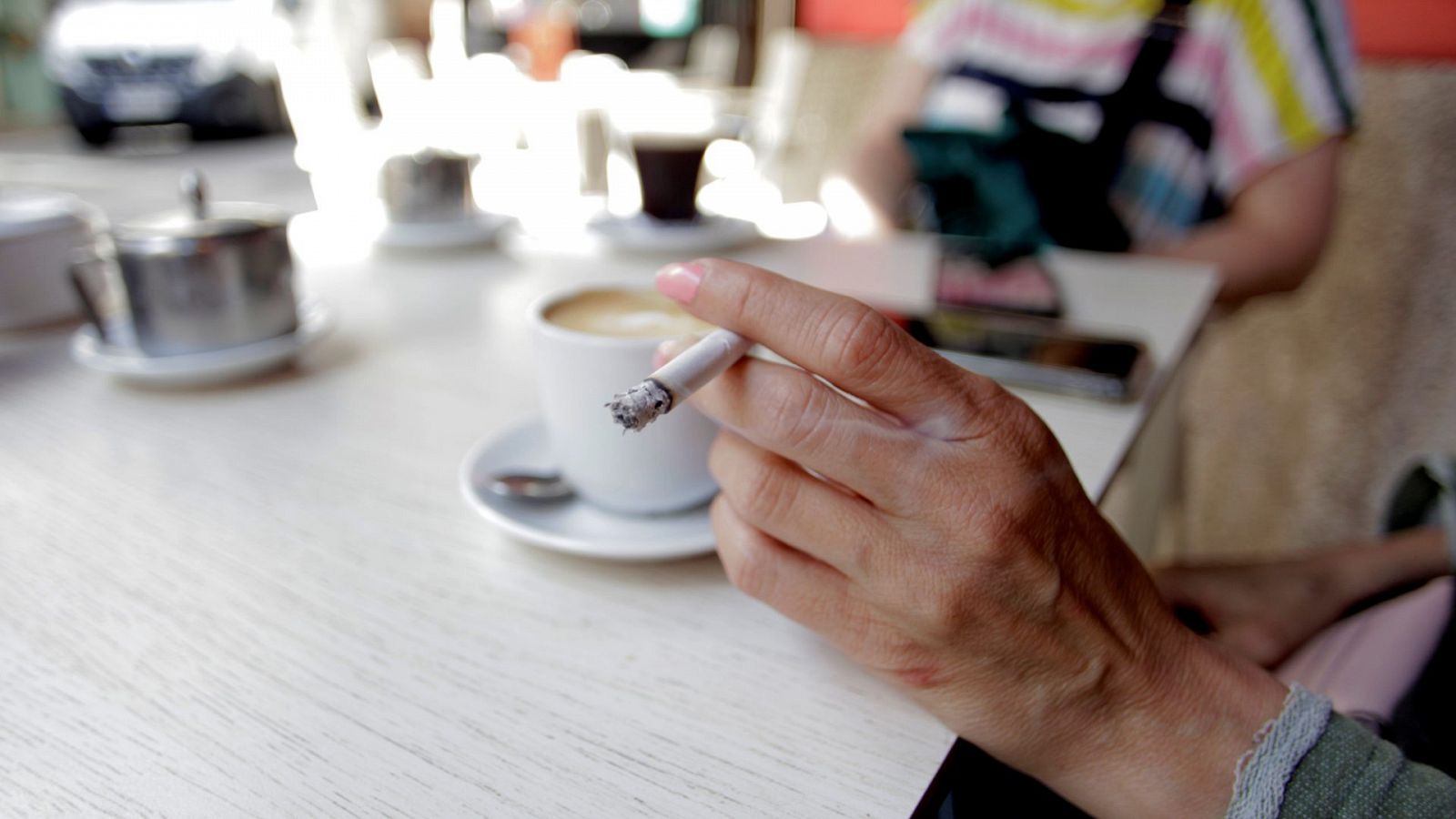 La mano d euna mujer sujeta un cigarro mientras se apoya en la mesa de una terraza de un bar. De fondo se ve diuminado un café